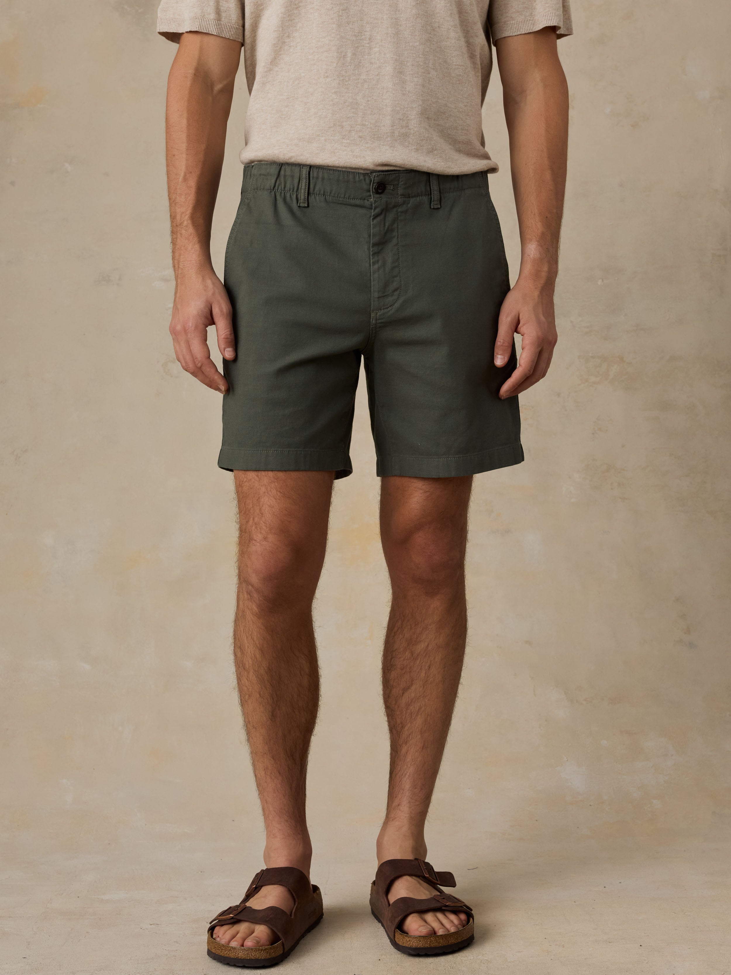 Man wearing dark olive green chino shorts with a beige t-shirt and sandals against a beige background