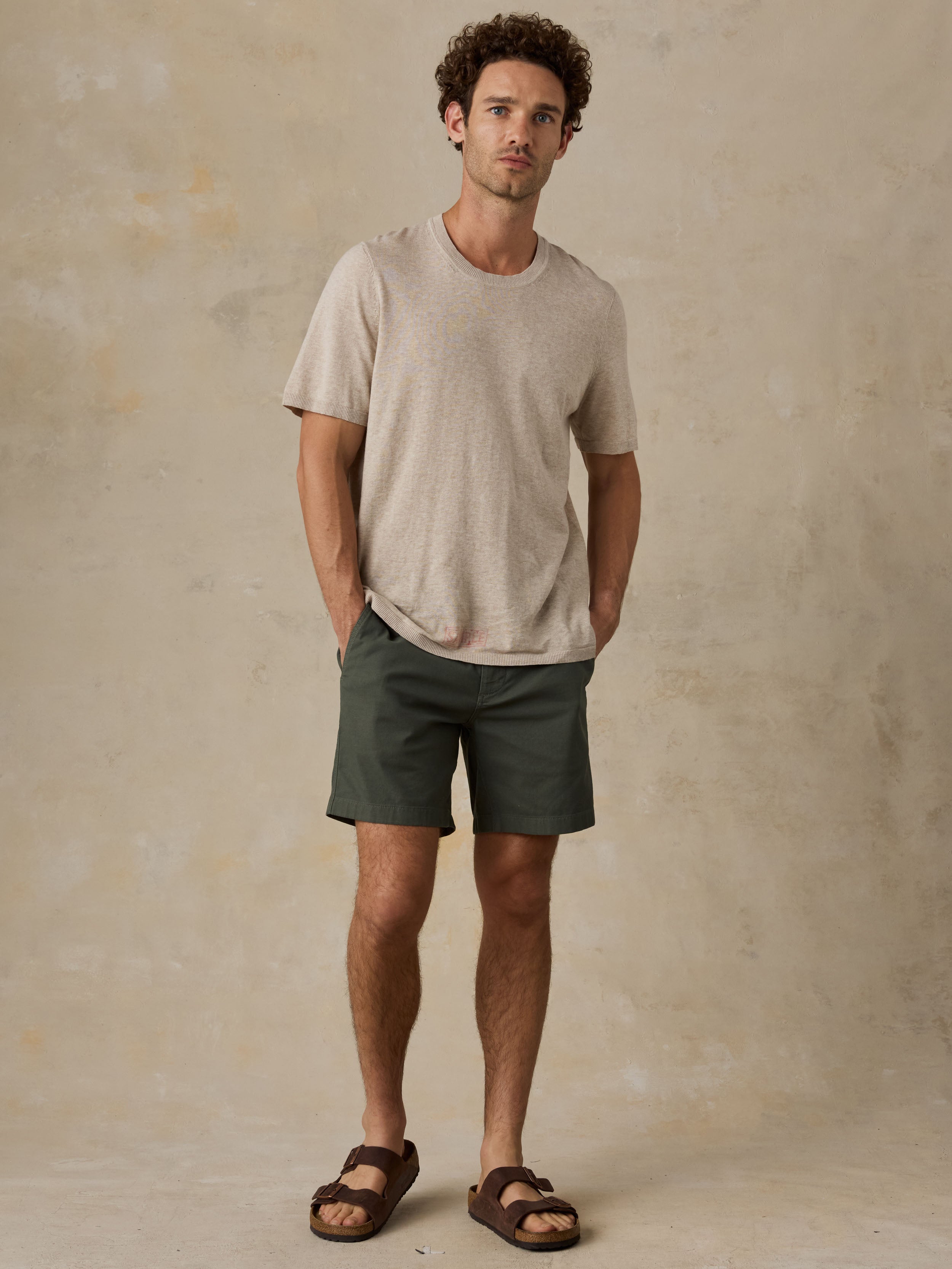 Man wearing dark olive green chino shorts with a beige t-shirt and sandals against a beige background