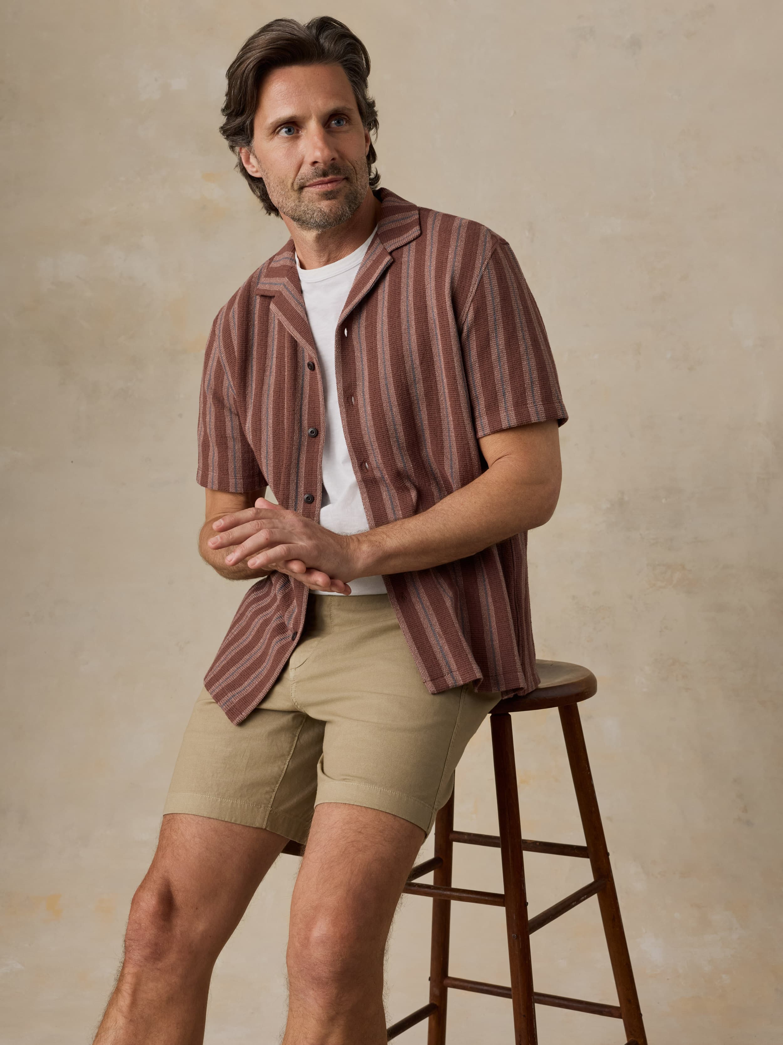 Man wearing a striped shirt and beige shorts sitting on a wooden stool against a beige wall.