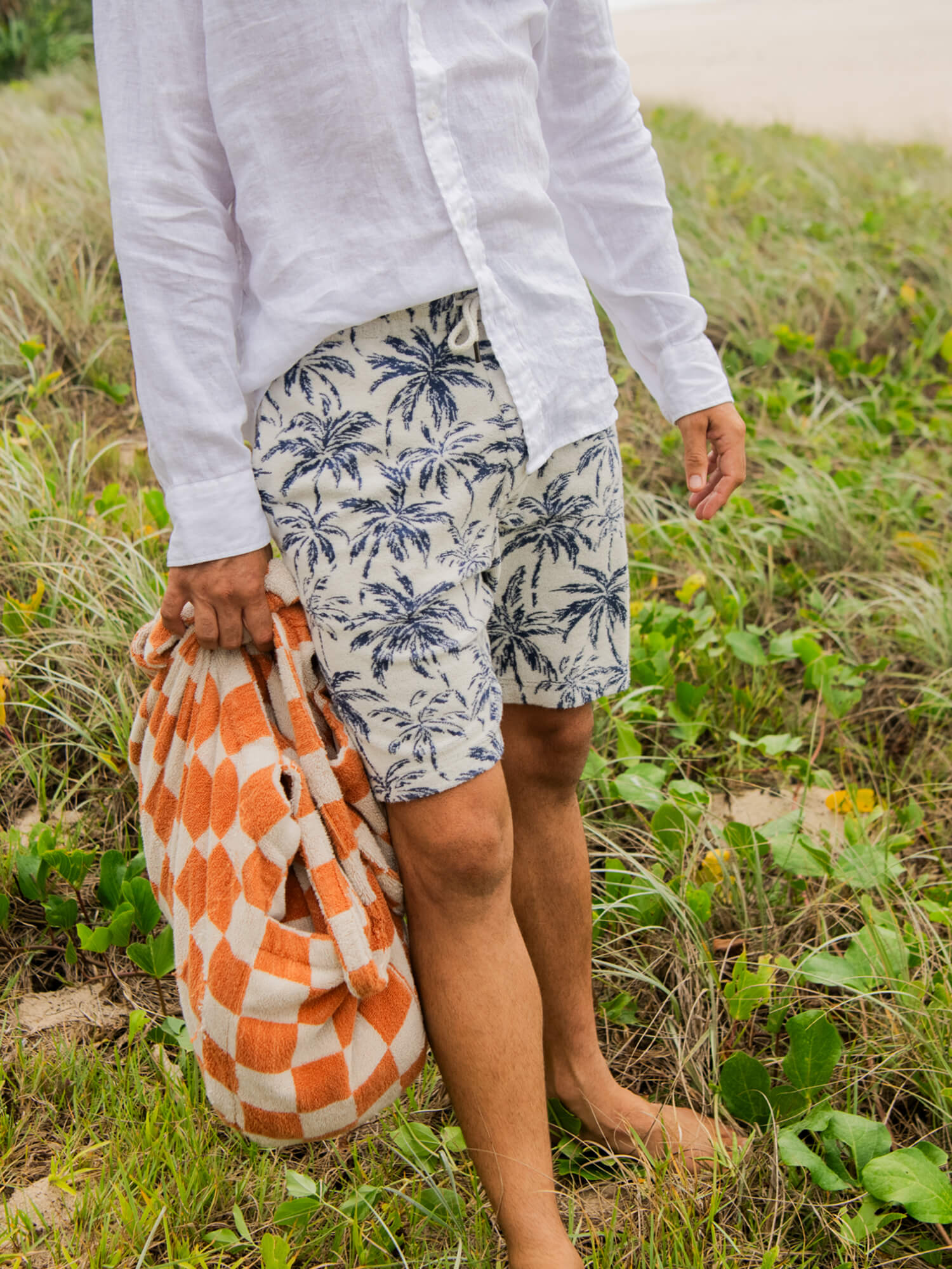 Man wearing palm tree printed shorts and a linen shirt standing in the grass holding a beach bag