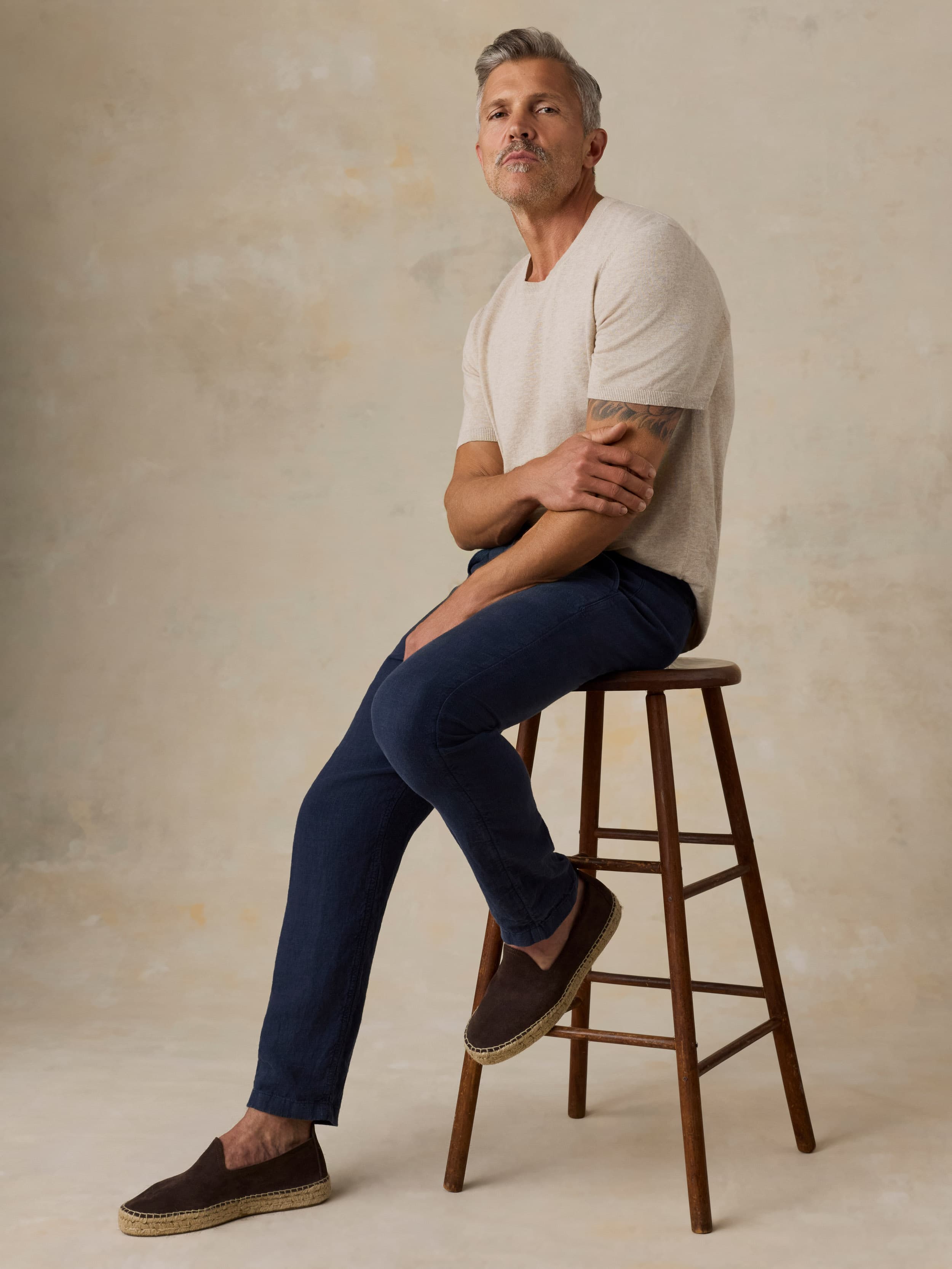 Man sitting on a wooden stool wearing navy linen pants and a beige t-shirt against a beige background