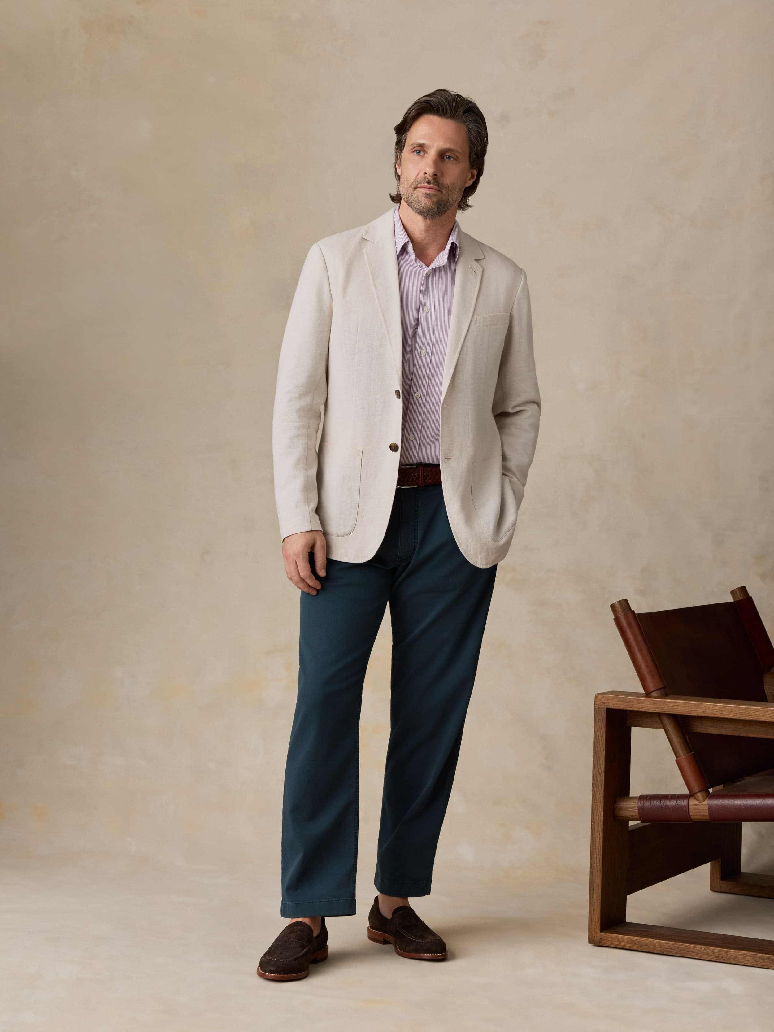 Man wearing navy pants with a purple button-down shirt and a light colored blazer against a beige background