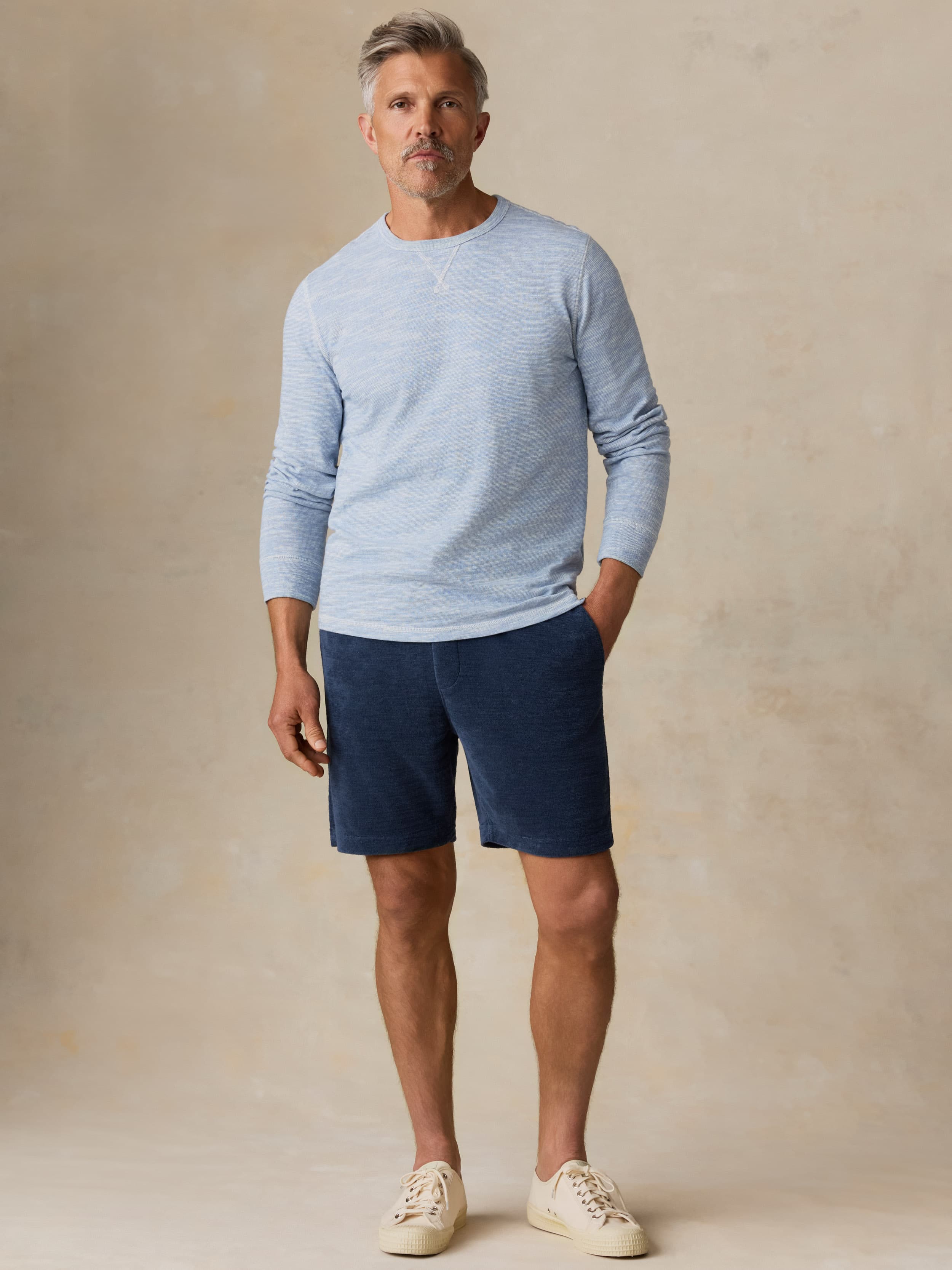 Man wearing a light blue crewneck and navy shorts against a beige background