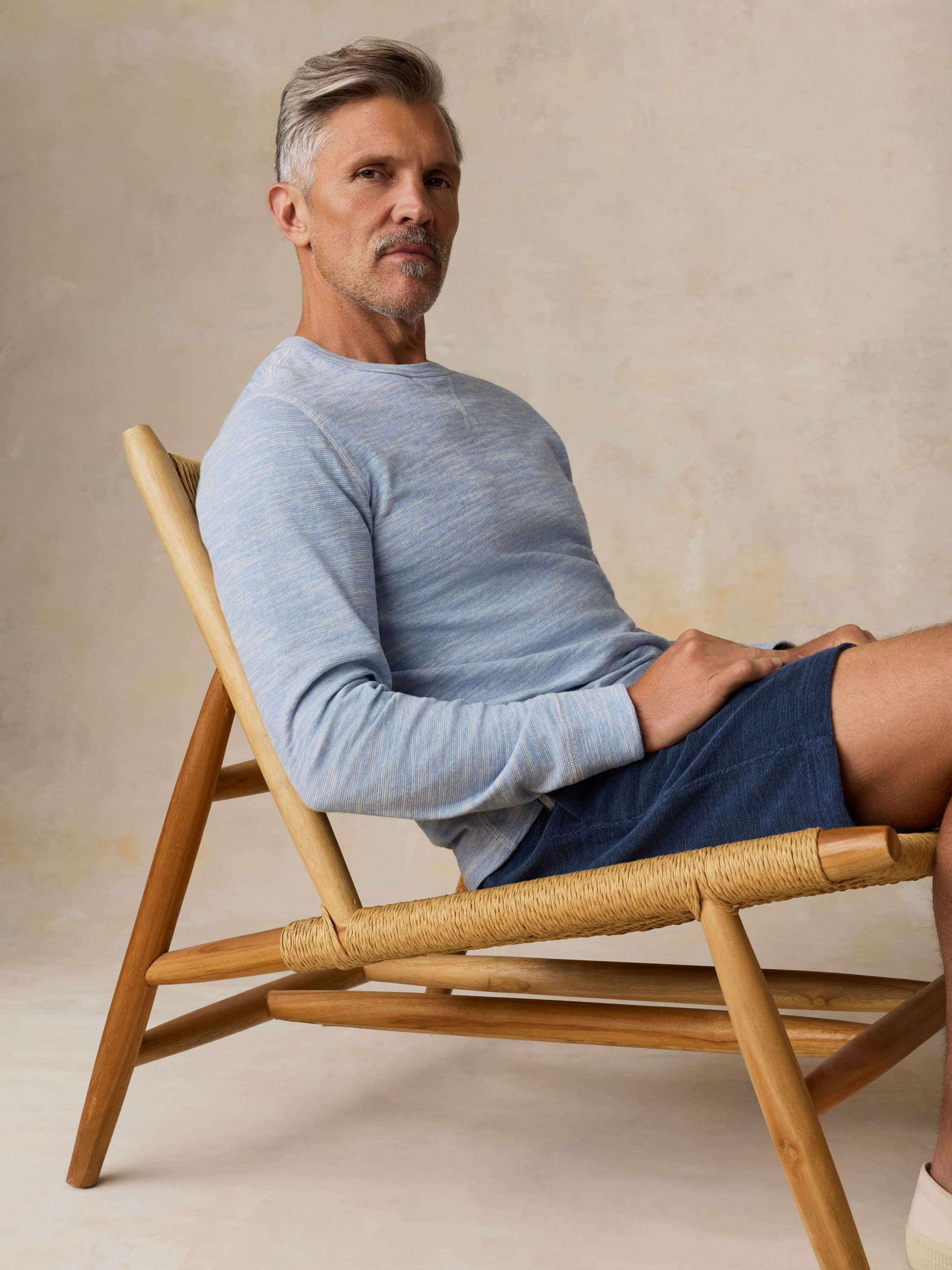 Man sitting on a wooden chair wearing a light blue crewneck and navy shorts against a beige background