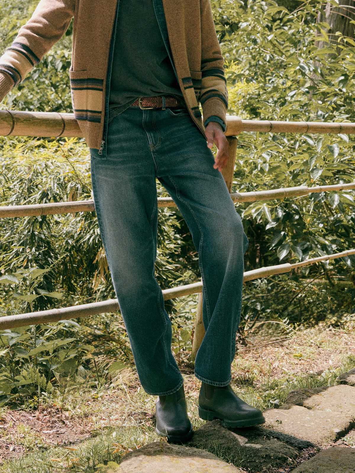 A man outdoors wearing blue denim jeans, a brown jacket, a dark green shirt, and dark rain boots, standing near a bamboo fence with green foliage in the background., image 1 of 7