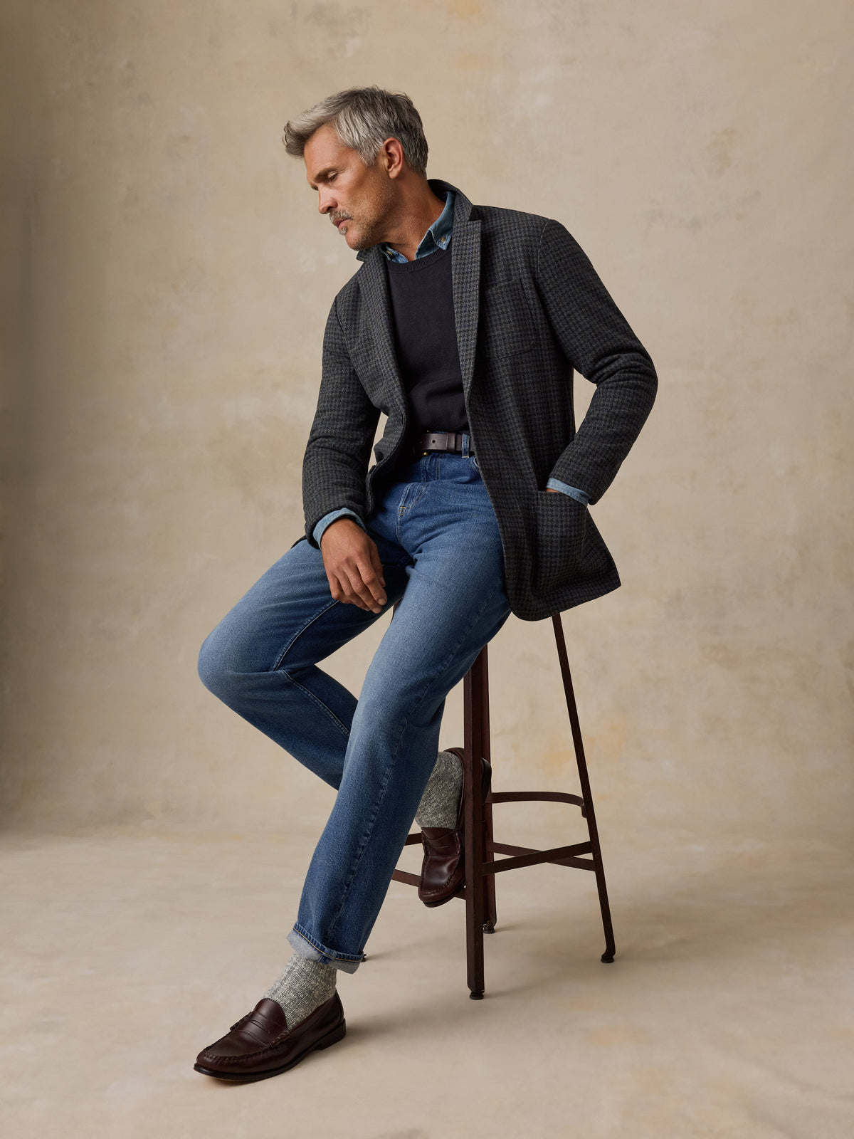 A man wearing a dark patterned blazer, blue jeans, a dark sweater, and brown loafers sits on a wooden stool against a textured beige studio backdrop., image 3 of 7