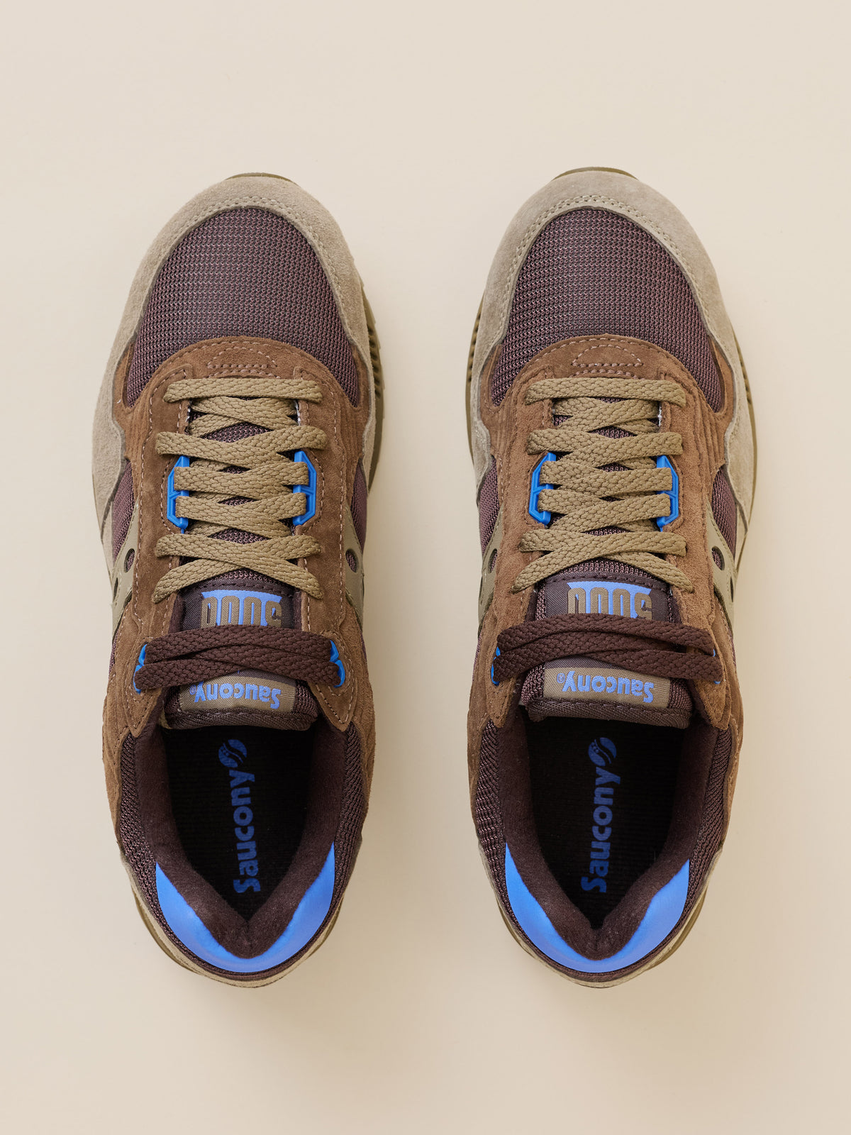 A pair of Saucony sneakers shown from a top-down view on a light beige background. The shoes feature a brown suede and mesh upper with dark purple overlays, beige laces, and bright blue accents on the tongue, heel, and inner sole., image 2 of 4