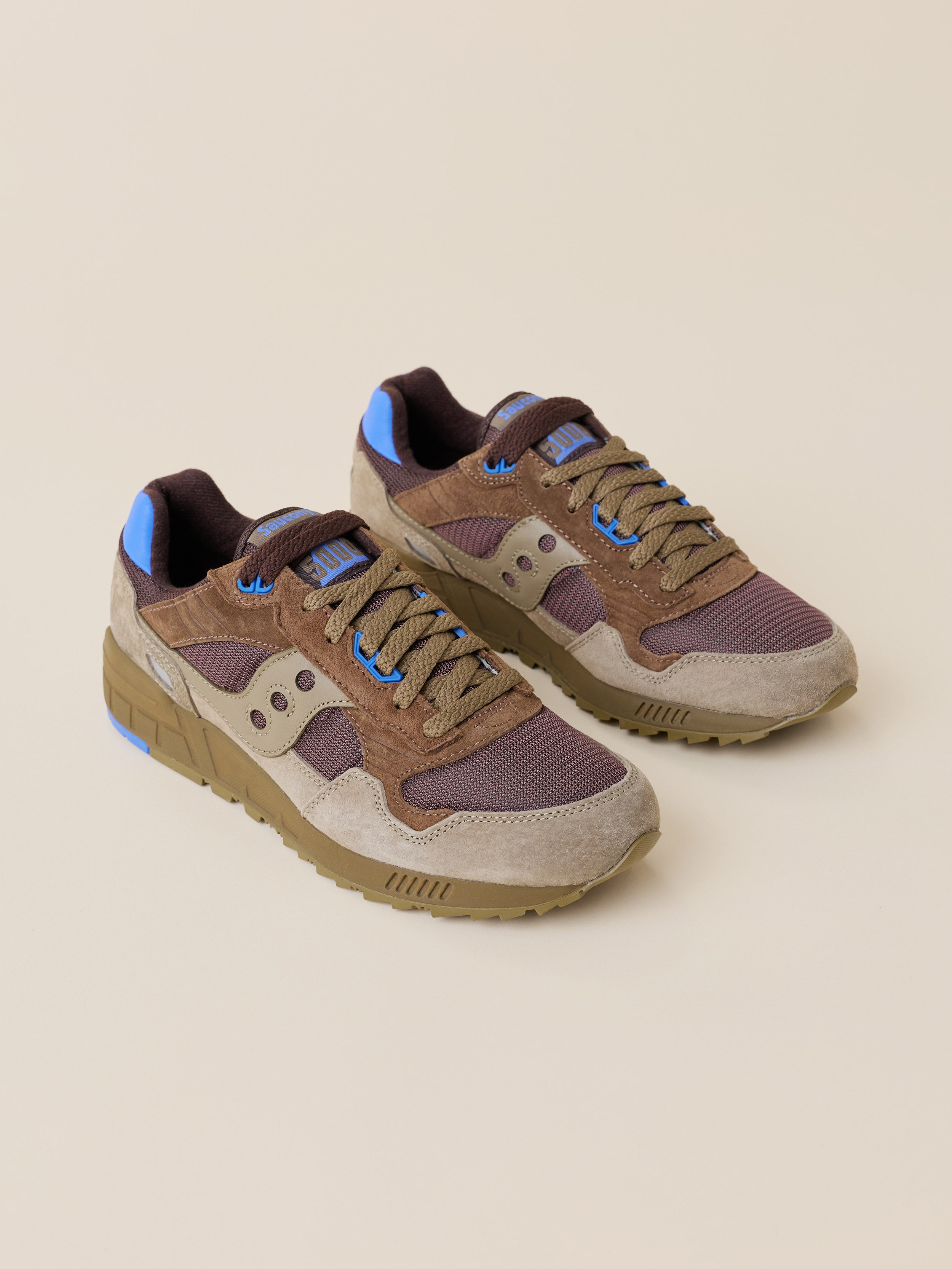 A pair of Saucony sneakers in brown, beige, and purple suede and mesh, featuring bright blue accents on the laces and tongue, set against a solid beige background.