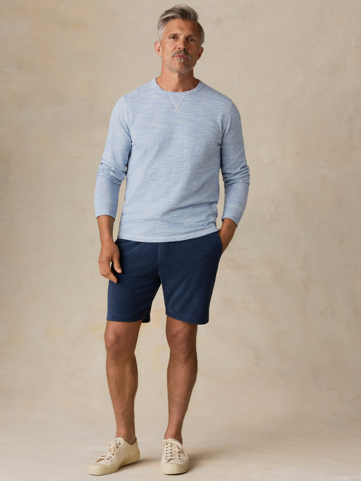 Man wearing a light blue crewneck and navy shorts against a beige background, image 3 of 7