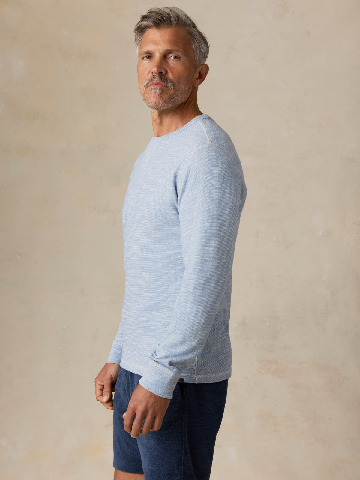 Man wearing a light blue crewneck and navy shorts against a beige background, image 4 of 7