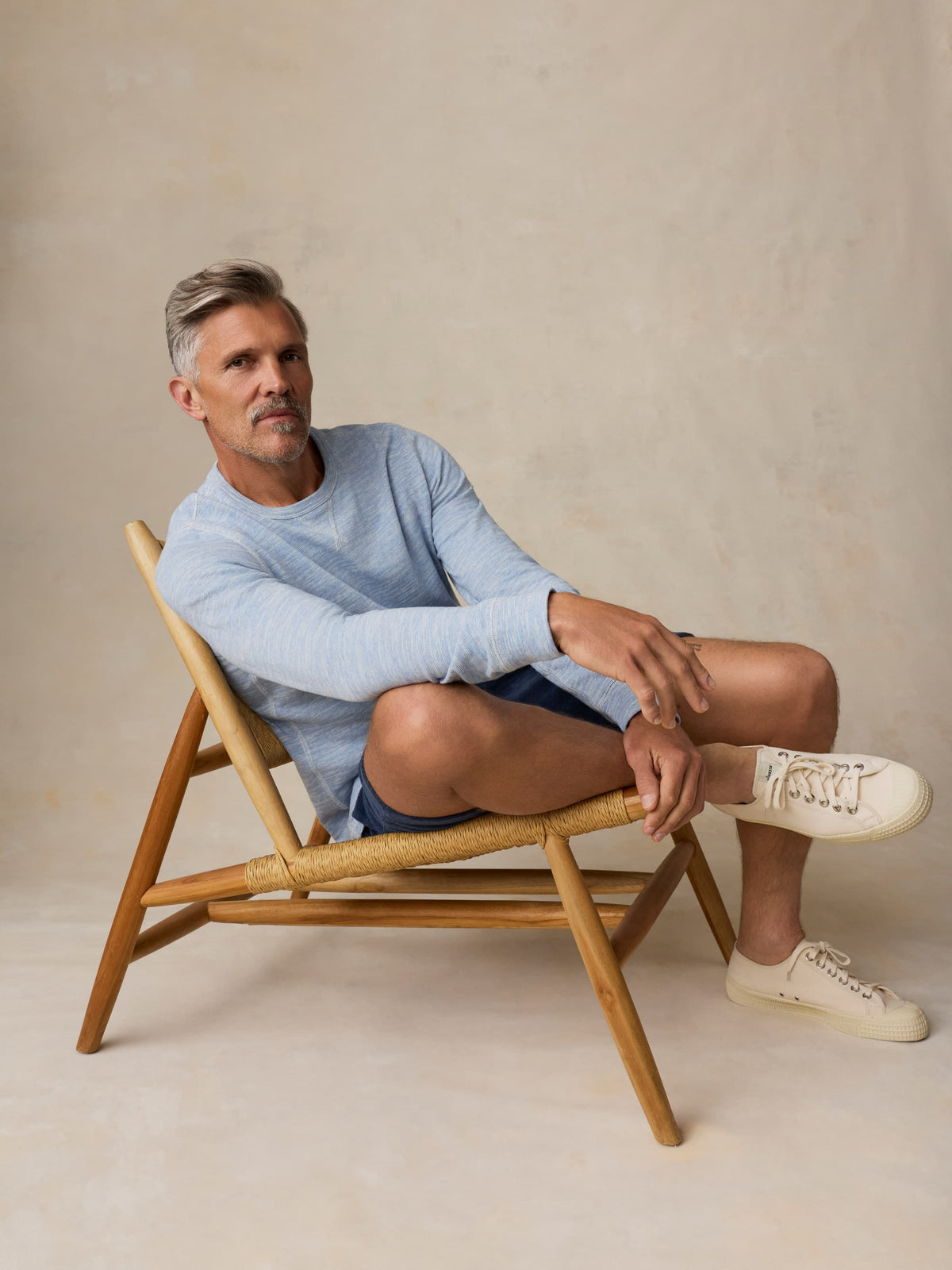 Man sitting on a wooden chair wearing a light blue crewneck and shorts against a beige background, image 2 of 7