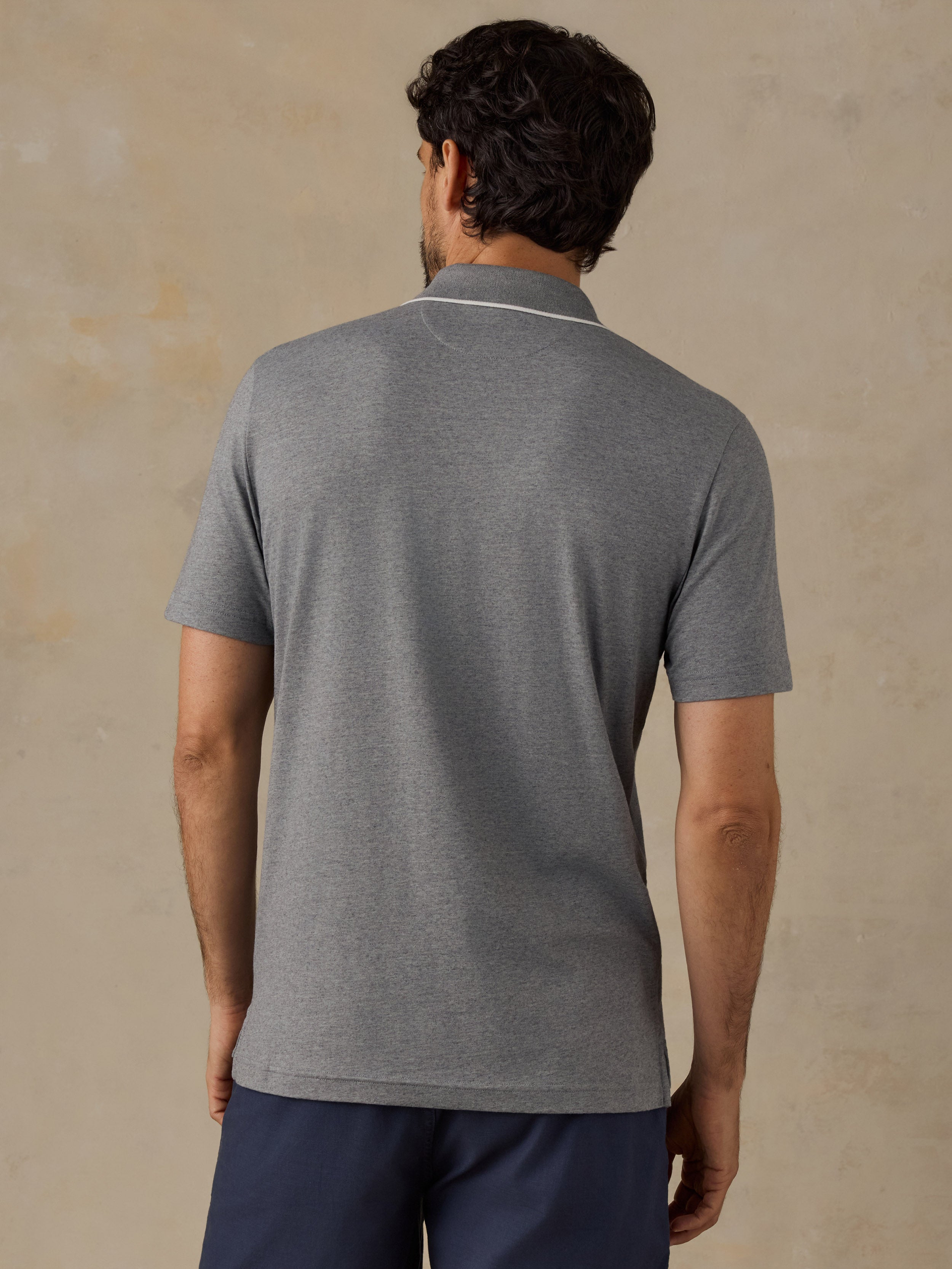 A man viewed from behind wearing a heather gray short-sleeve polo shirt with a thin white stripe detail at the collar, paired with navy blue pants, set against a textured beige background.