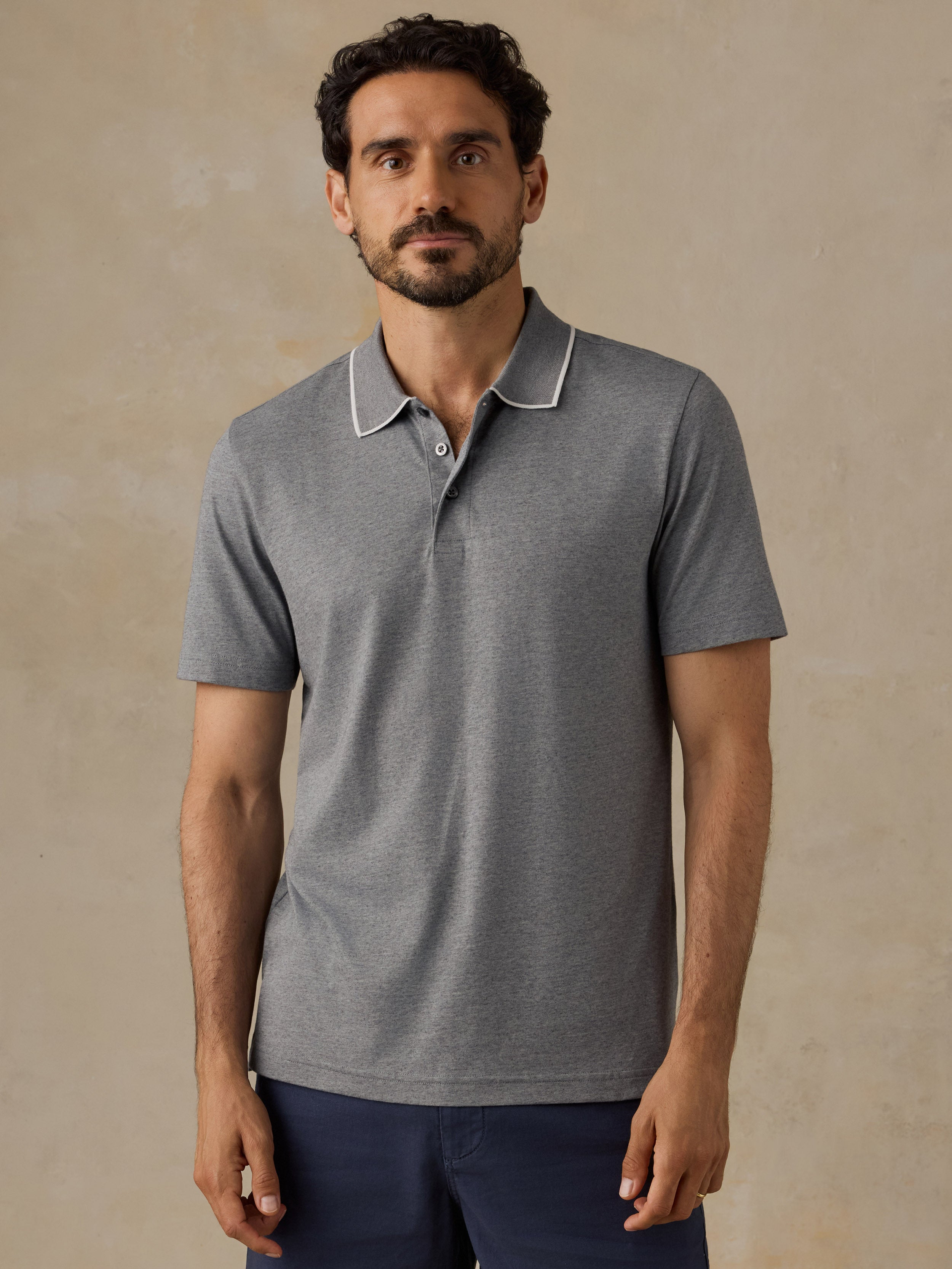 A man wearing a heather gray short-sleeve polo shirt with a white contrast collar, paired with navy blue pants, standing against a textured beige background.