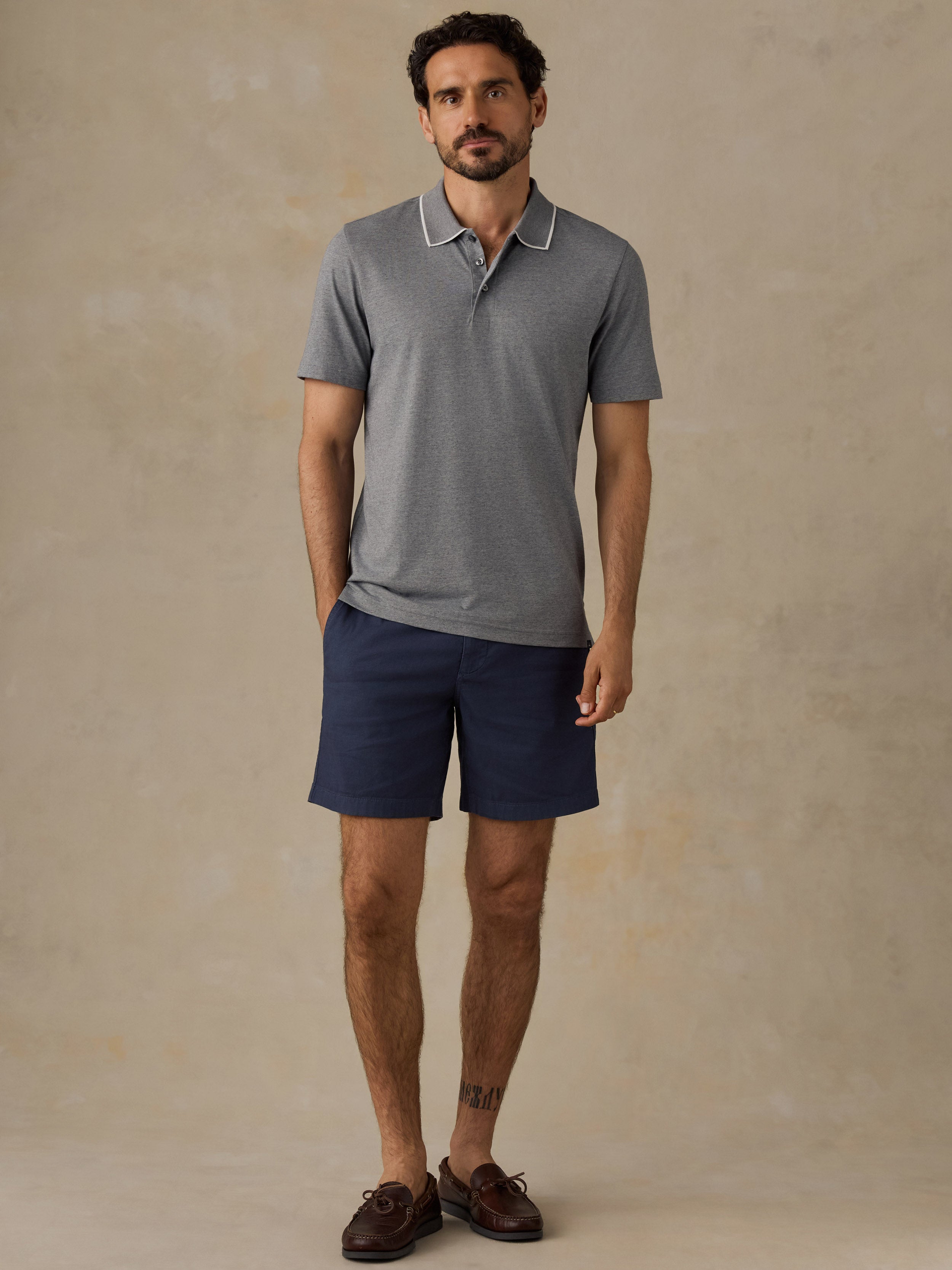 A man modeling a short-sleeve polo shirt in heather gray with a white contrast collar, paired with navy blue shorts and brown boat shoes, standing against a textured beige studio backdrop.
