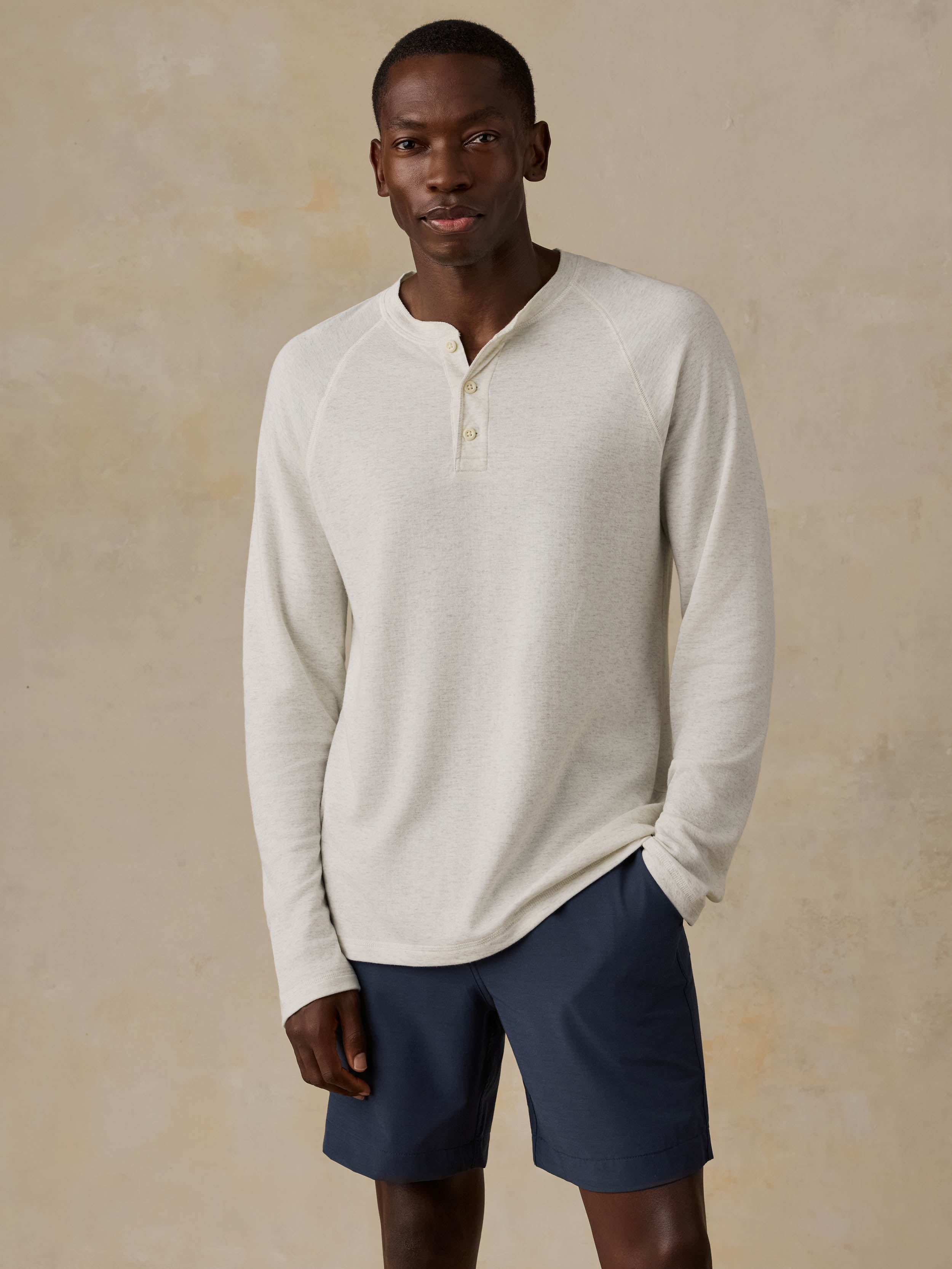 Man is wearing a cream colored long-sleeve henley shirt and navy shorts against a beige background