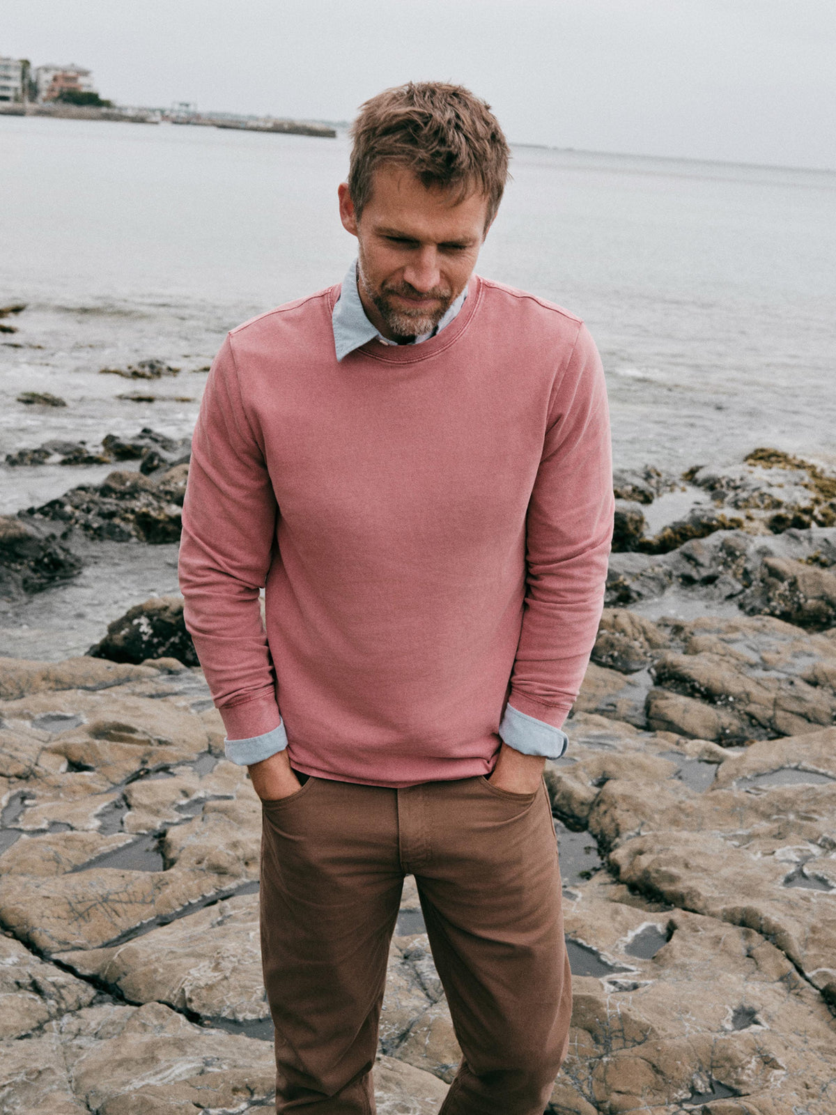 A man standing on rocky shoreline wearing a dusty rose long-sleeve crewneck sweater over a light blue collared shirt, paired with brown trousers., image 1 of 7