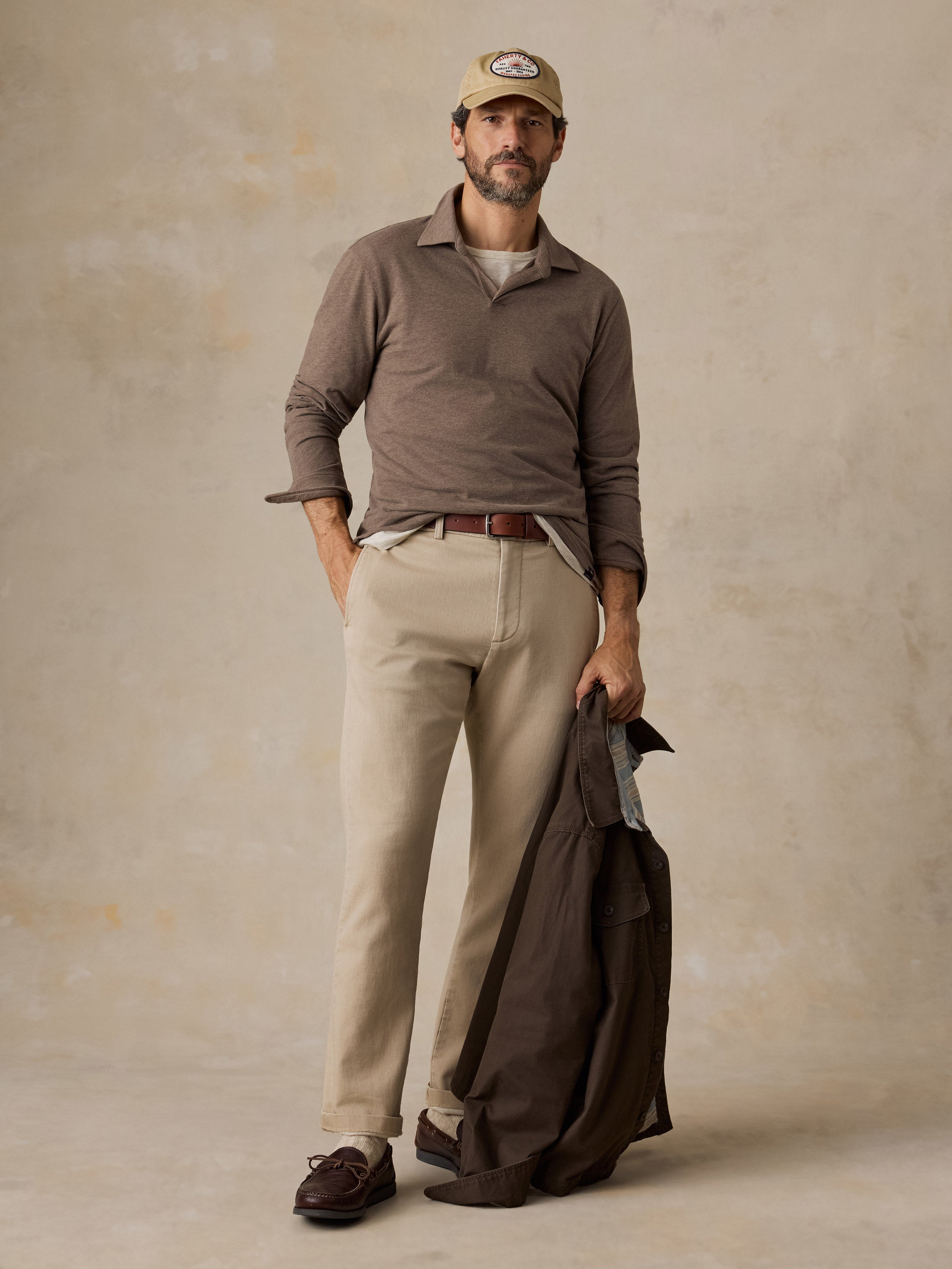 A man wearing a brown polo shirt, beige chino pants, beige hat, and brown jacket