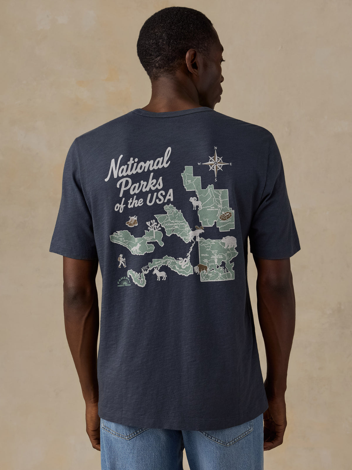 A man wearing a navy blue t-shirt with a large back graphic of a map of National Parks of the USA, shown from the back against a textured beige background., image 2 of 7