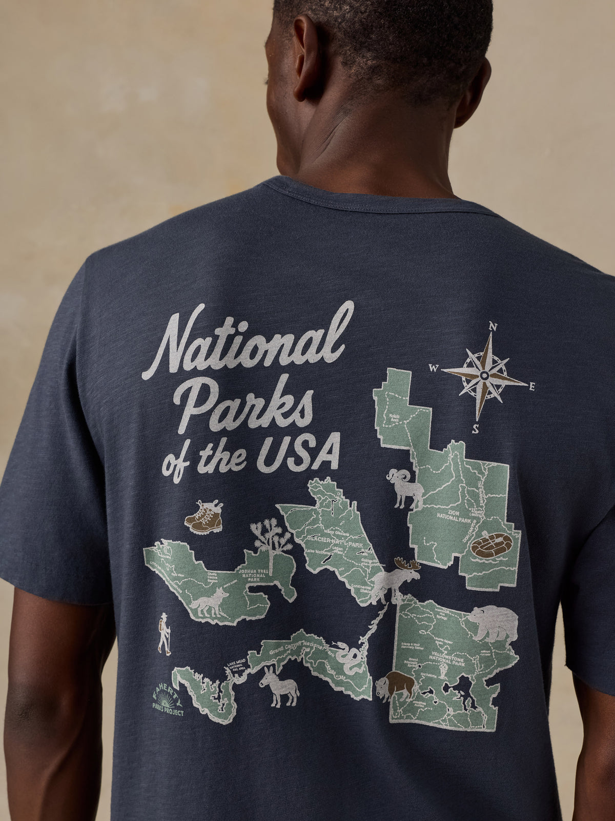 A man viewed from behind wearing a navy blue t-shirt with a large graphic print on the back. The graphic features a map of National Parks of the USA in light green and white, complete with a compass rose and various animal illustrations., image 5 of 7