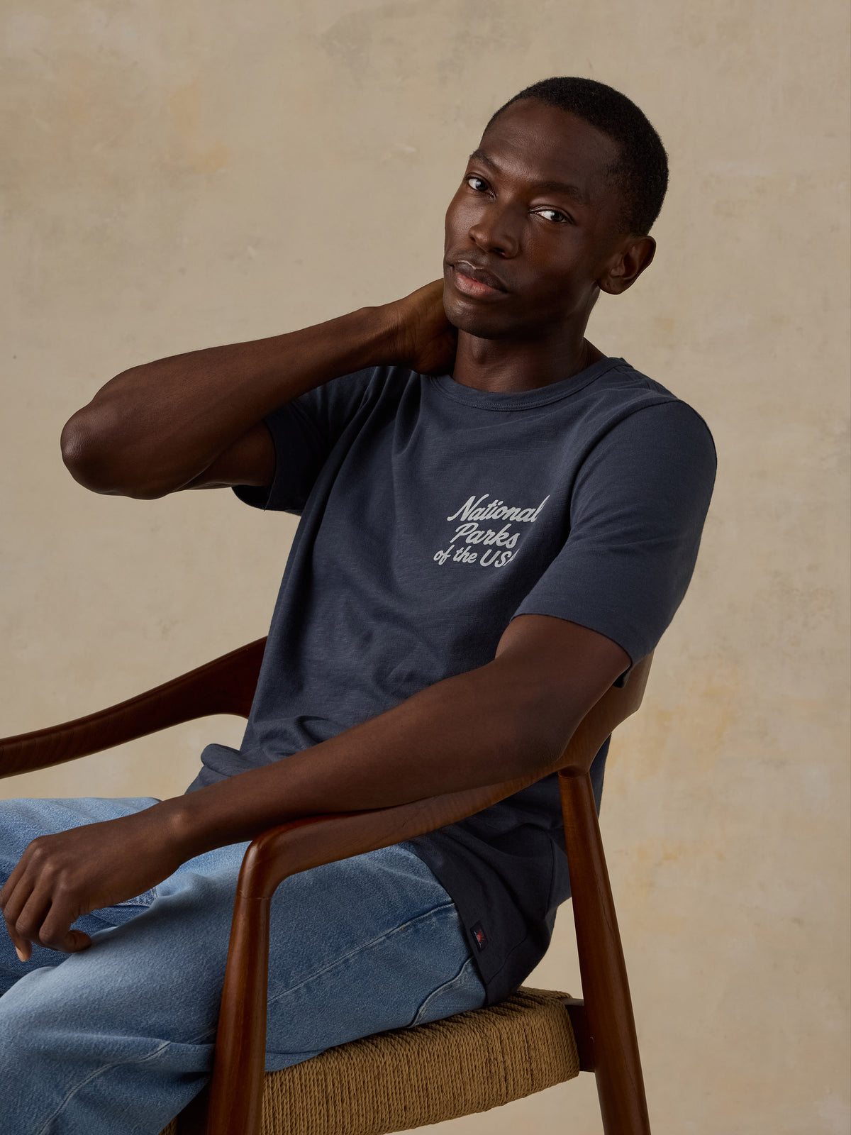 A man wearing a navy blue t-shirt with white cursive text reading 'National Parks of the US' sits in a wooden armchair against a textured beige background., image 3 of 7