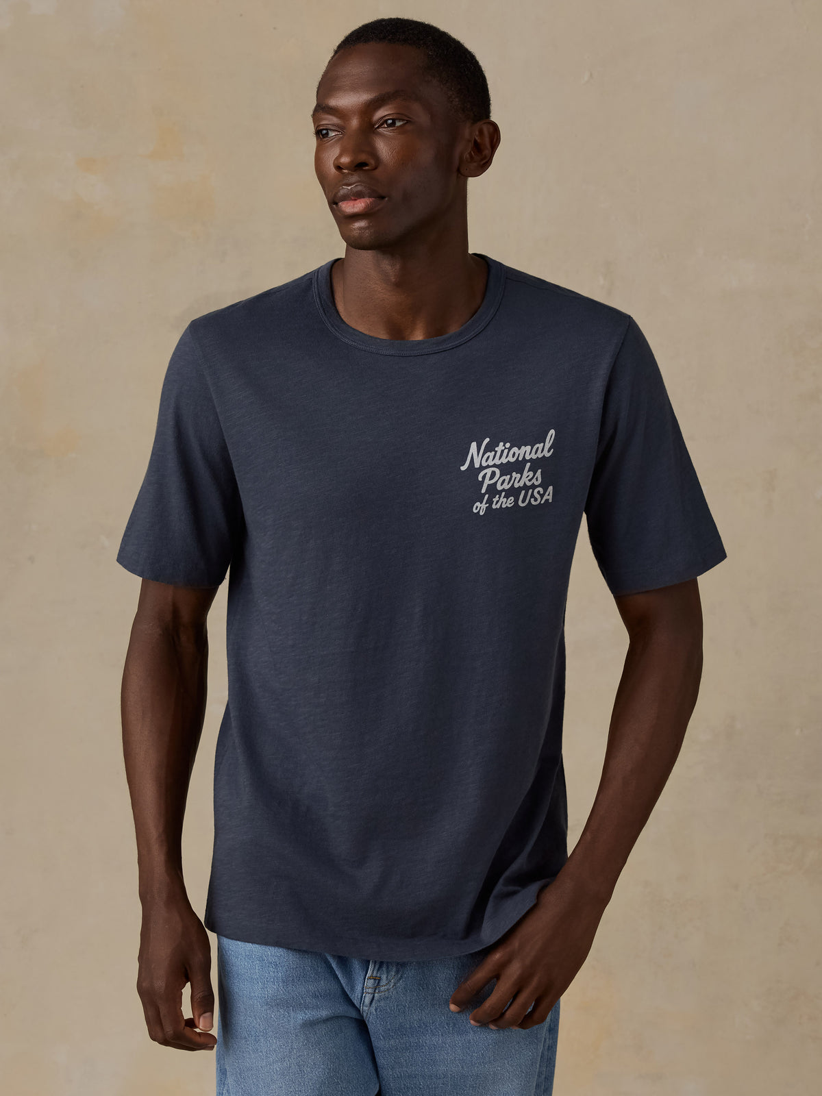 A man wearing a navy blue crewneck t-shirt with 'National Parks of the USA' printed in white script on the left chest, paired with light blue jeans, set against a textured beige background., image 1 of 7