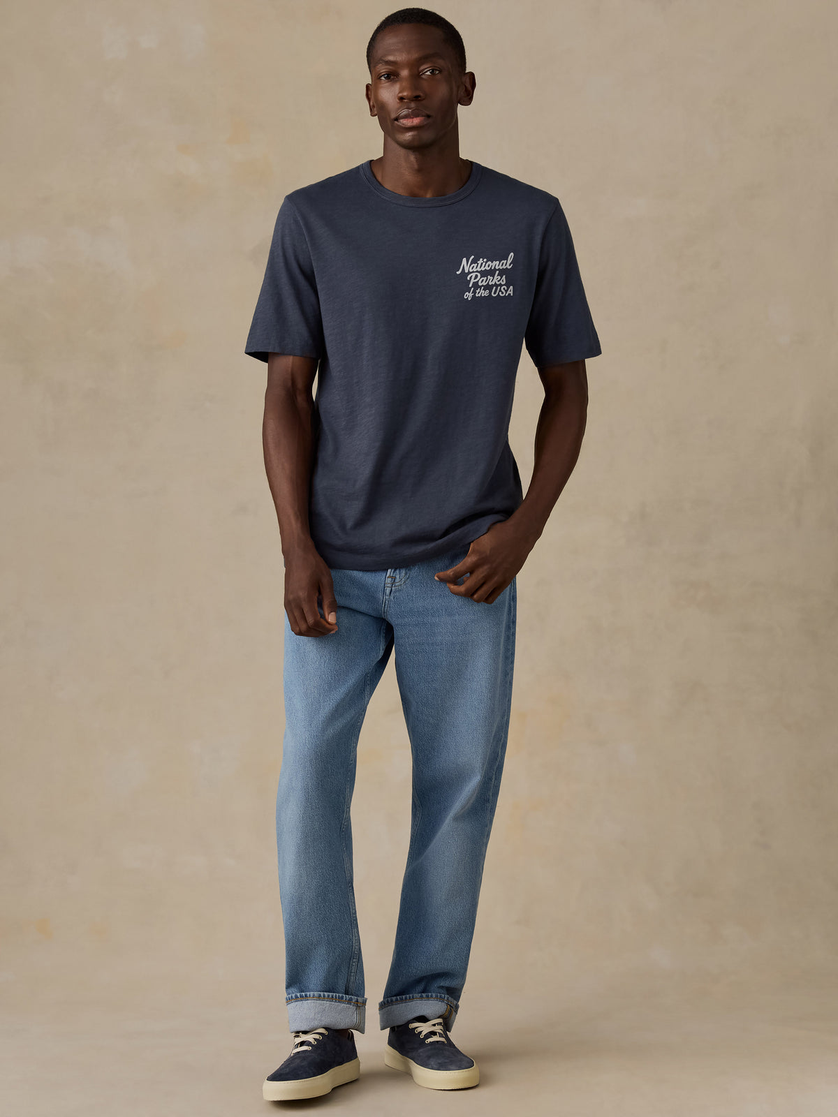 A man modeling a navy blue crewneck t-shirt with a white 'National Parks of the USA' graphic on the left chest, paired with light blue jeans and navy sneakers, against a textured beige studio backdrop., image 4 of 7