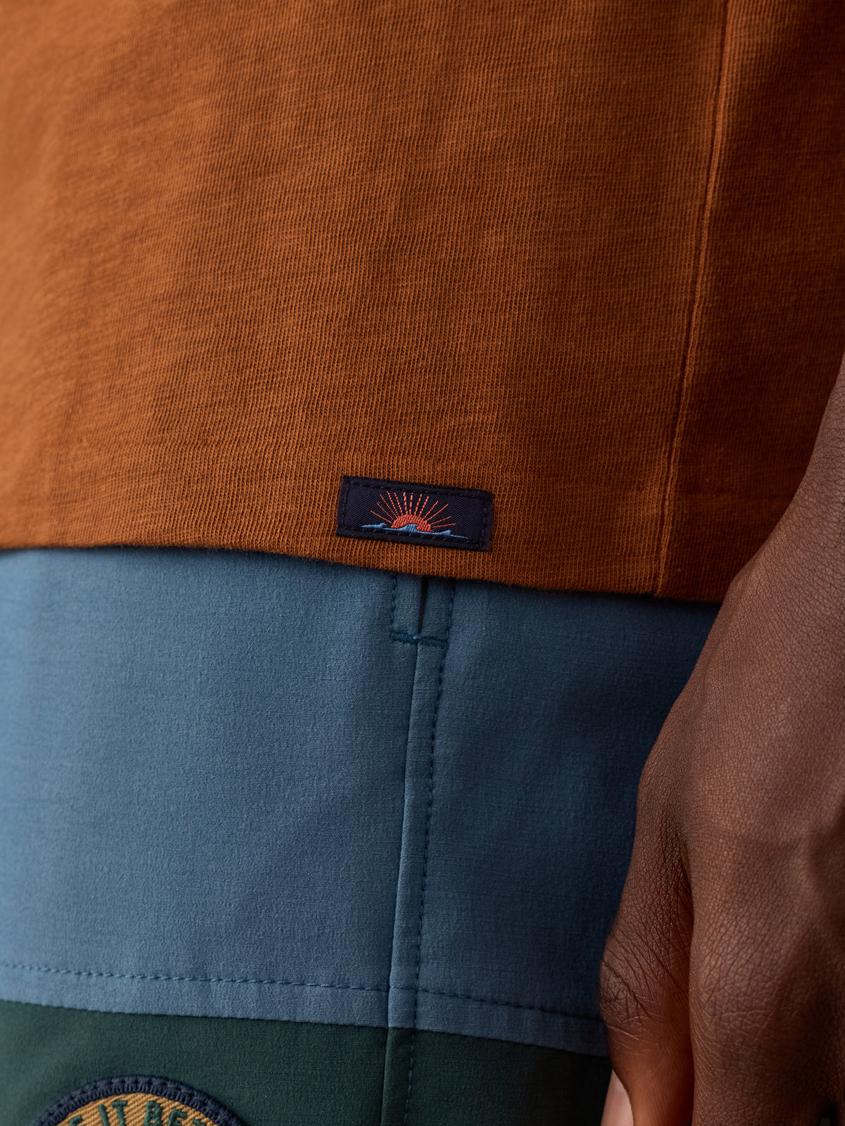 Close-up of a man wearing a rust-orange knit top layered over a blue and green color-blocked bottom, with a small Faherty brand logo patch near the hem., image 5 of 8