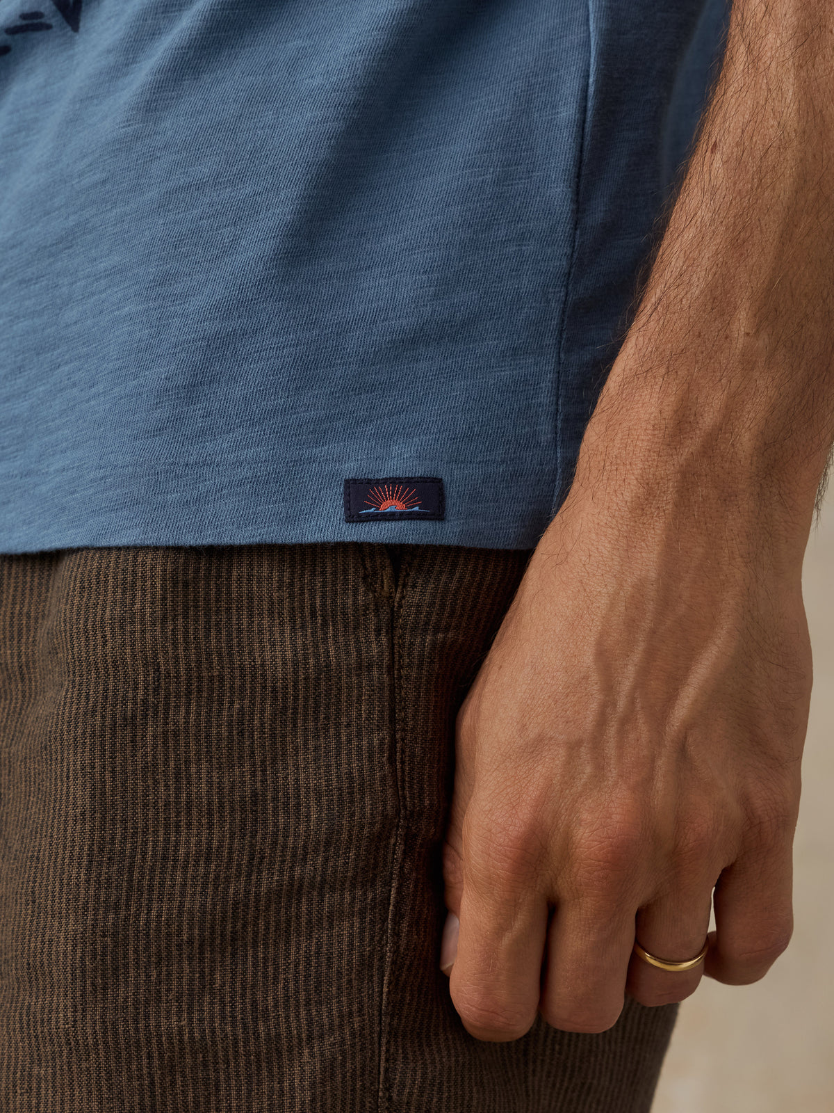 Close-up of a man wearing a heathered slate blue t-shirt and brown corduroy pants, with a small Faherty brand patch on the shirt hem., image 4 of 6