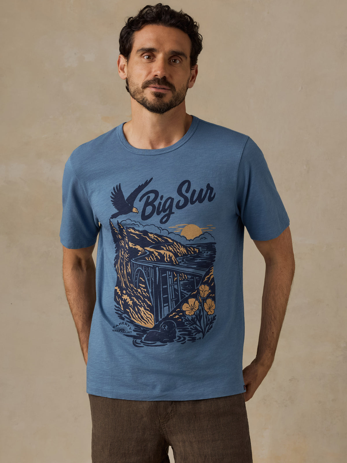 A man wearing a heather blue graphic t-shirt with a 'Big Sur' coastal illustration in navy and yellow, paired with brown pants, standing against a textured beige wall., image 1 of 6