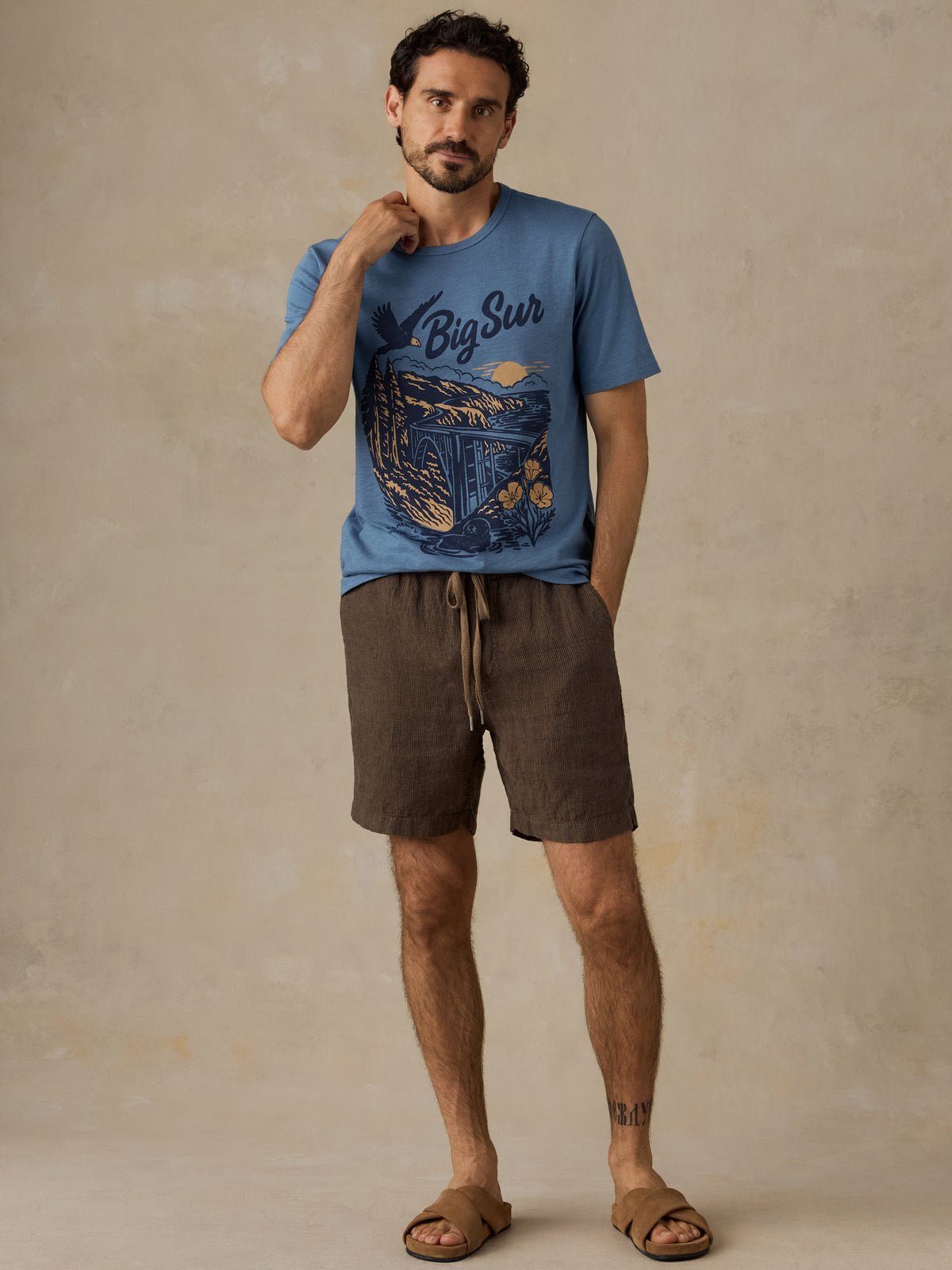 A man modeling a blue graphic t-shirt with 'Big Sur' written on it, paired with brown drawstring shorts and brown leather slide sandals, standing against a textured beige studio backdrop., image 2 of 6