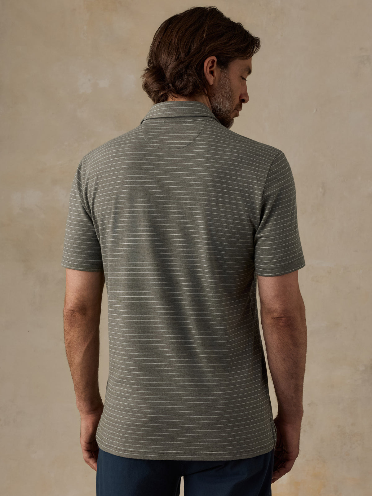 A man viewed from the back wearing a short-sleeve polo shirt with fine horizontal stripes in olive green and gray, paired with dark blue pants, set against a textured beige background., image 3 of 5