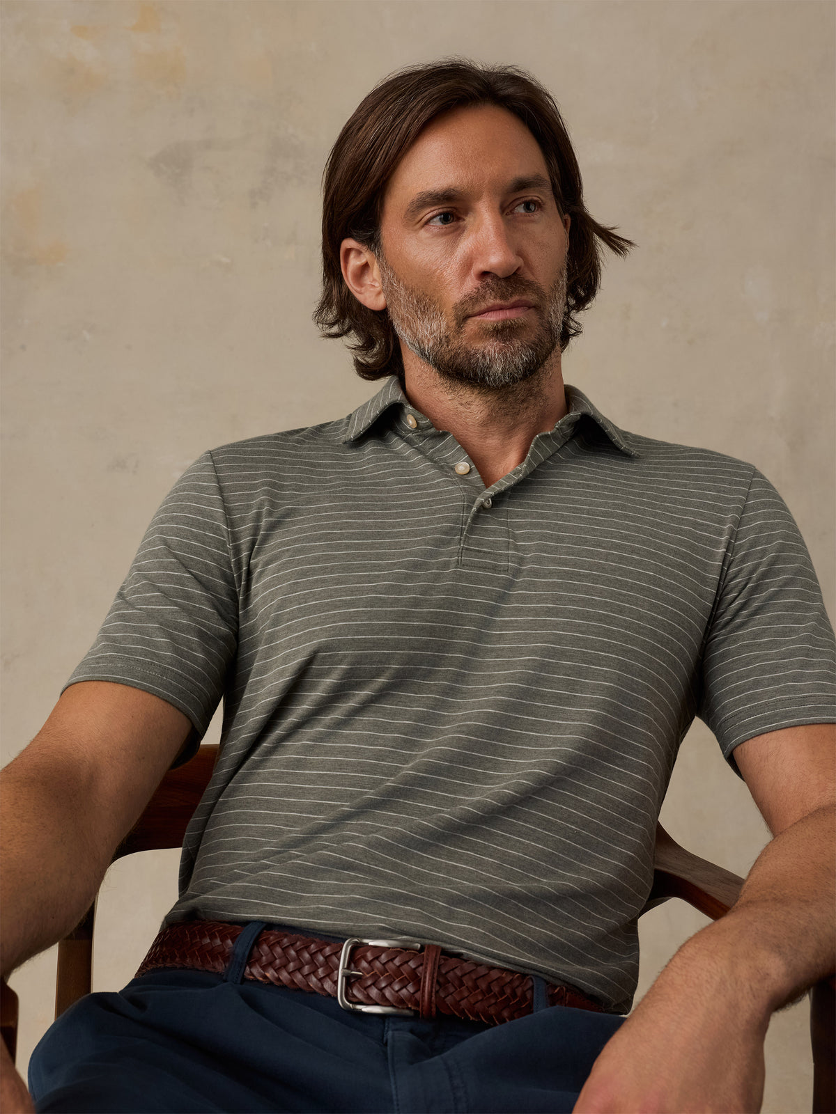 A man wearing a short-sleeve polo shirt with thin horizontal gray stripes on an olive green base, paired with a brown braided belt and navy pants, seated in a wooden chair against a textured beige wall., image 5 of 5