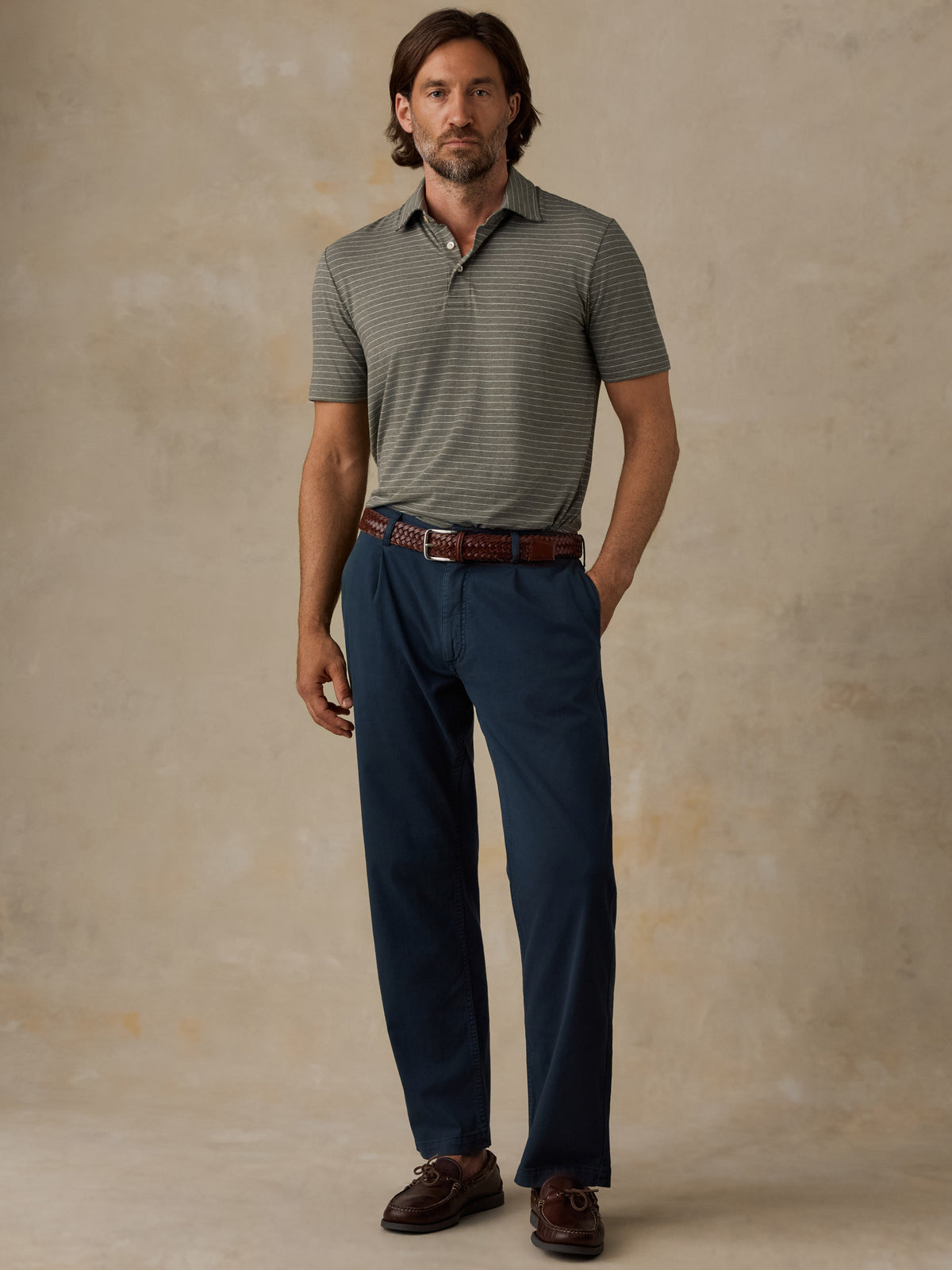A man modeling a short-sleeve polo shirt with fine horizontal stripes in gray and olive green, paired with navy blue trousers, a brown leather belt, and brown loafers, standing against a textured beige studio backdrop., image 4 of 5
