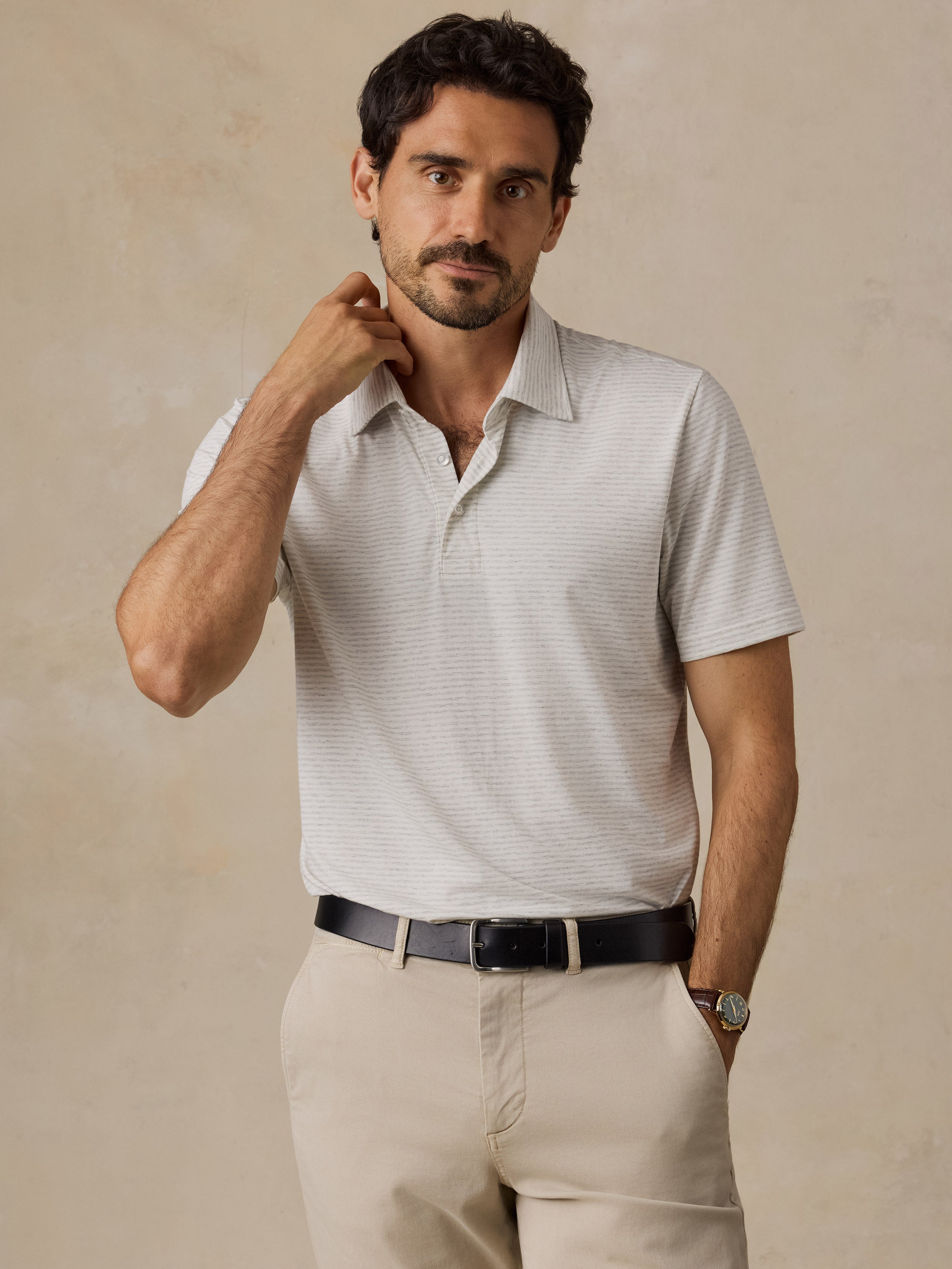 A man wearing a light beige short-sleeve polo shirt with a subtle textured pattern, paired with beige trousers and a black belt, posing against a neutral beige background.
