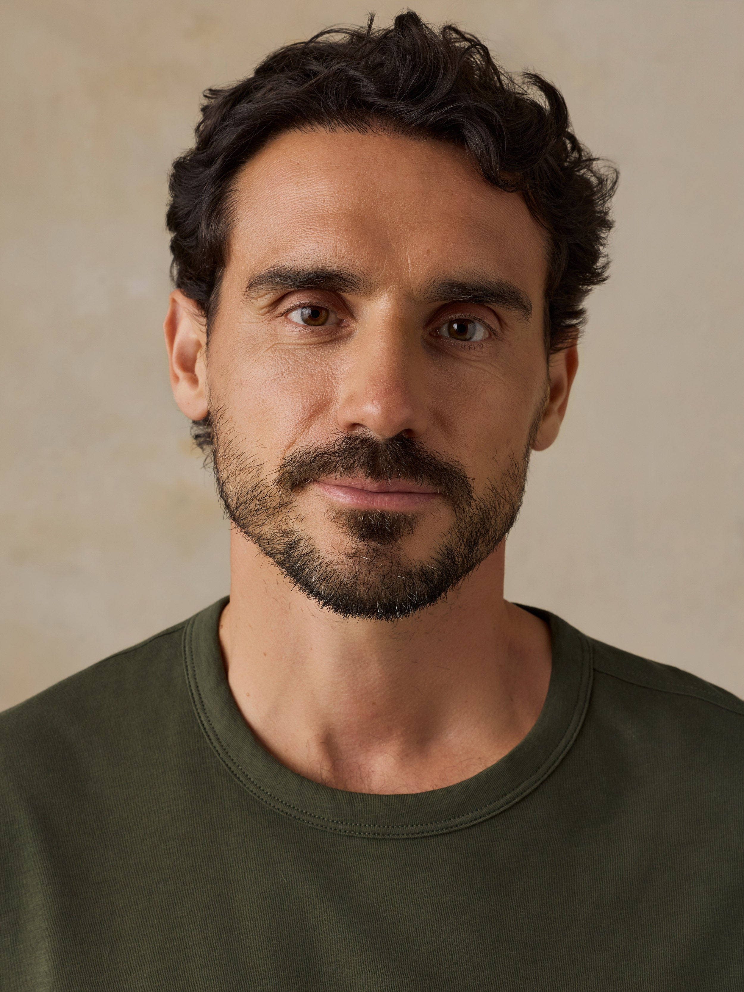 A close-up portrait of a man with dark curly hair, brown eyes, and a short beard, wearing an olive green crew-neck t-shirt against a neutral beige background.