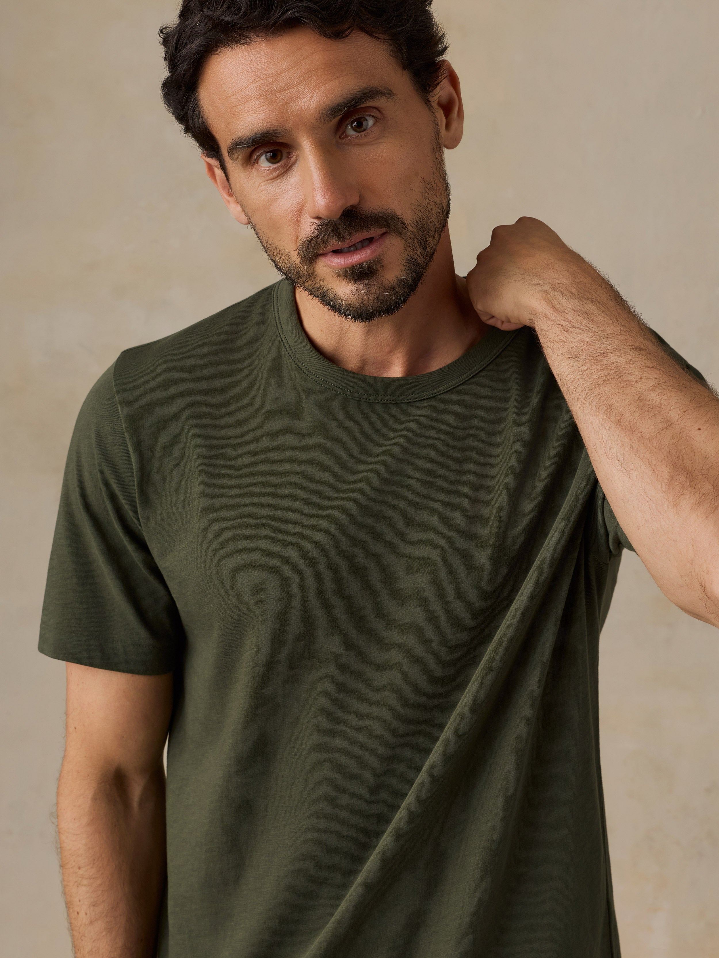 A man wearing a solid olive green short-sleeve crewneck t-shirt, posing with one hand on his neck against a neutral beige textured background.