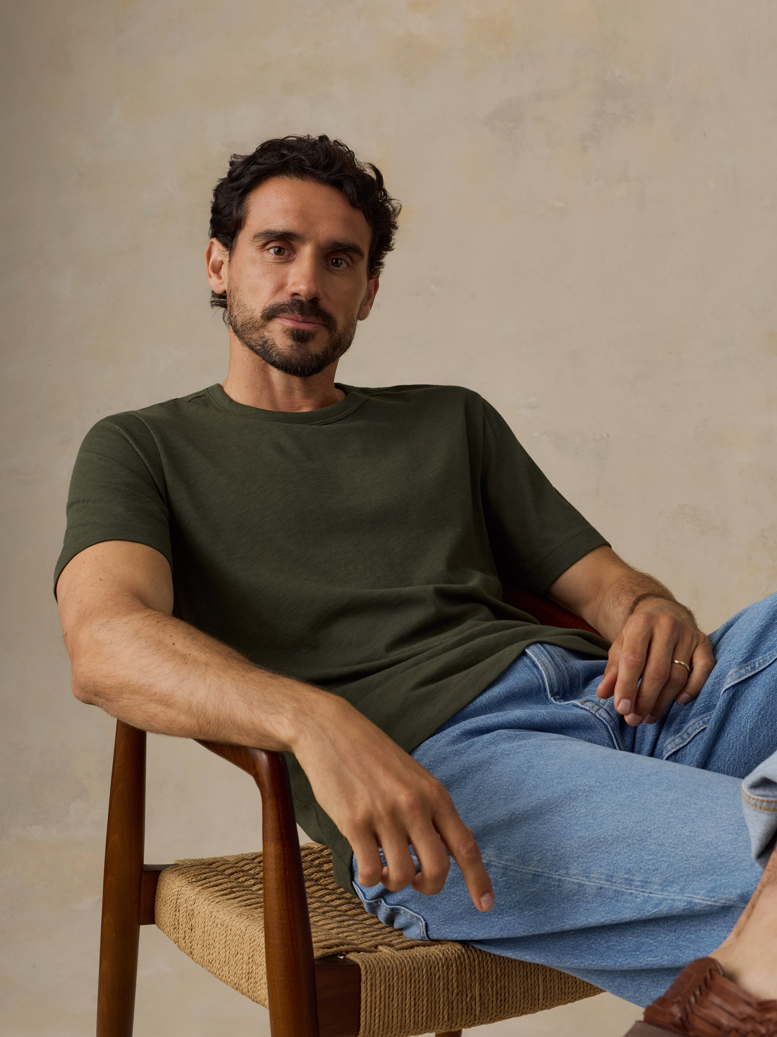 A man wearing an olive green crew-neck t-shirt and light blue jeans, sitting in a wooden chair with a woven seat against a textured beige wall.