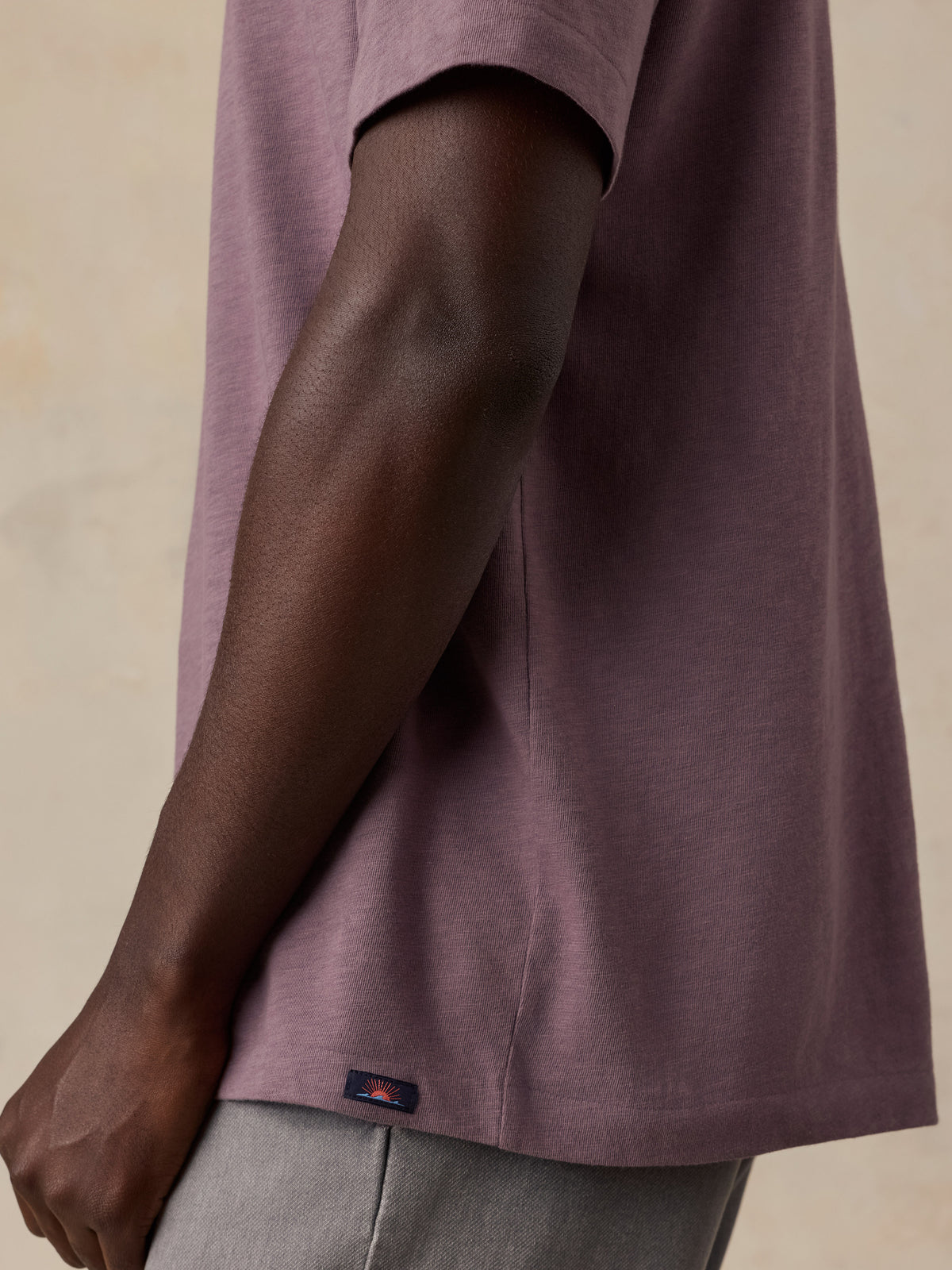 Close-up side view of a man wearing a muted mauve-purple short-sleeve T-shirt and light gray pants, with a small dark Faherty brand tab near the hem., image 6 of 6