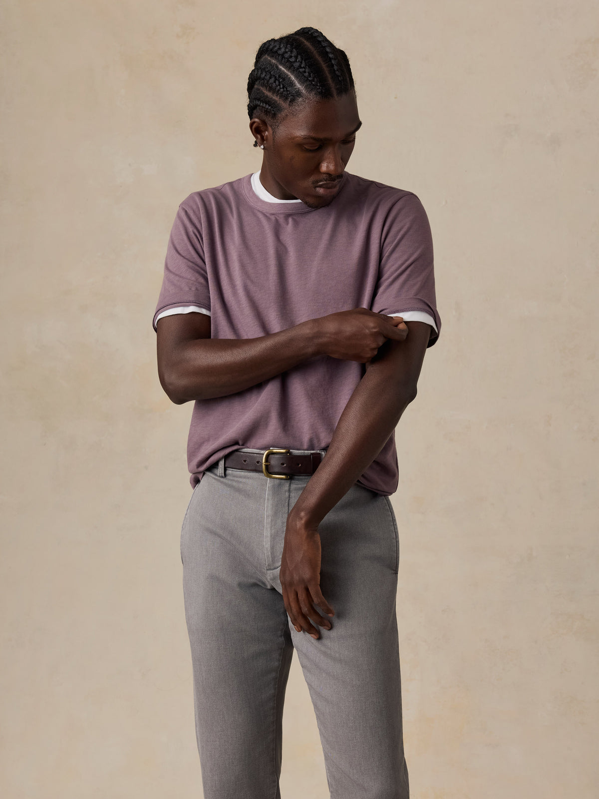 A man wearing a mauve-purple t-shirt paired with gray trousers and a dark belt, standing against a textured beige background., image 4 of 6