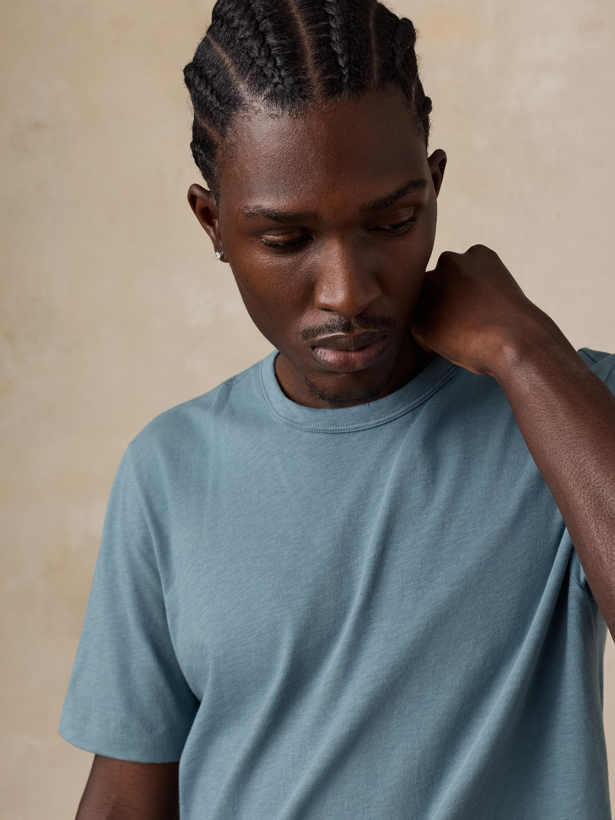 A man with braided hair wearing a muted blue-gray crewneck t-shirt, posing with his hand on his neck against a textured beige background., image 2 of 5