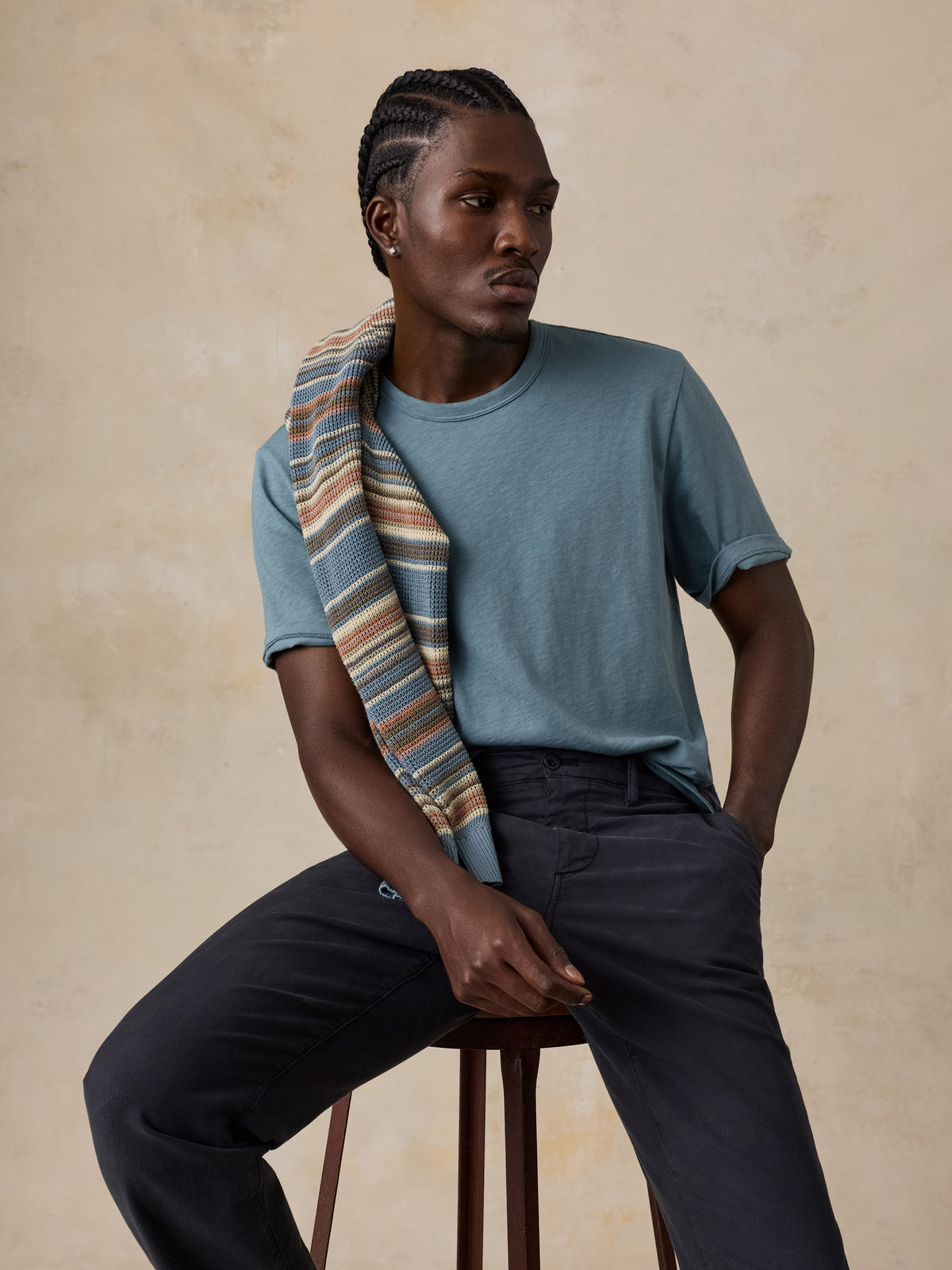 A man seated on a stool wearing a muted blue crew-neck t-shirt, dark charcoal trousers, and a striped scarf draped over his shoulder, posed against a textured beige studio backdrop., image 3 of 5
