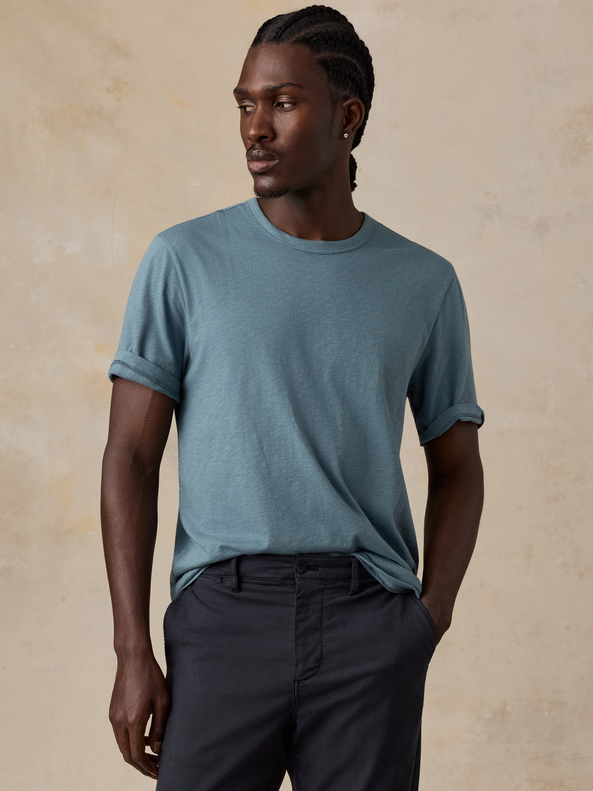 A man wearing a muted blue-gray crewneck t-shirt with rolled sleeves and dark charcoal pants, standing against a textured beige studio backdrop.