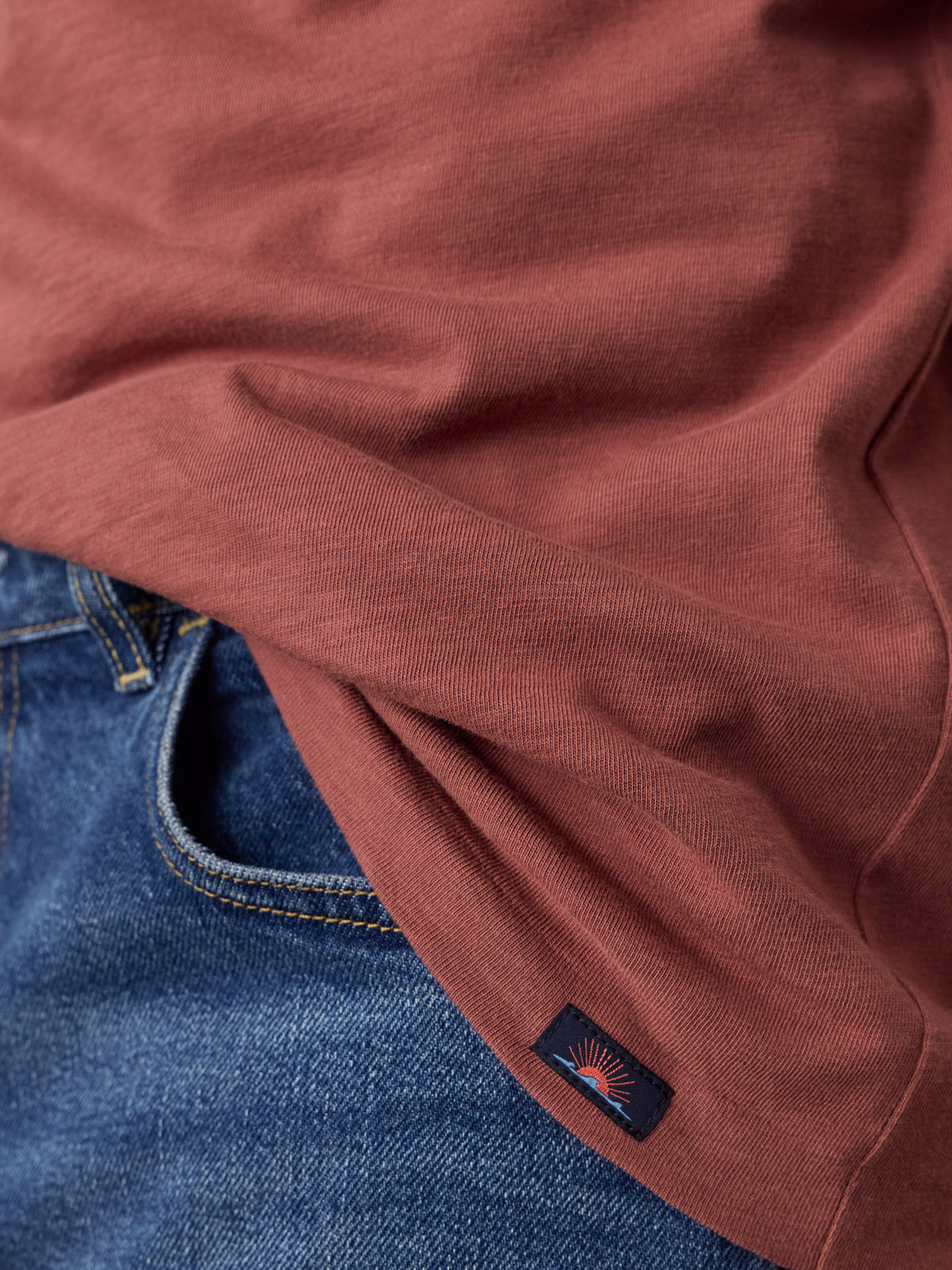 Close-up of a rust-colored t-shirt draped over blue denim jeans, showing a small Faherty brand patch near the hem, image 4 of 6