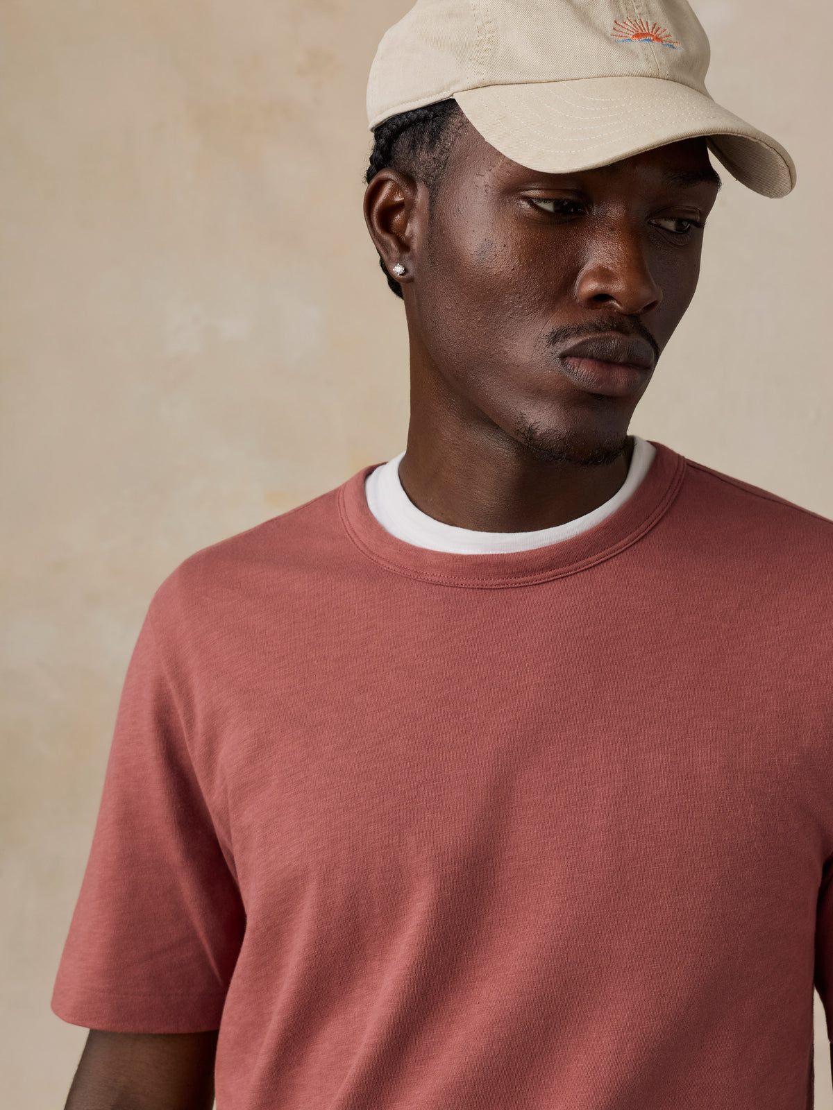 A man wearing a dusty rose crewneck t-shirt with a white ribbed collar and a beige baseball cap, photographed against a textured beige background., image 5 of 6