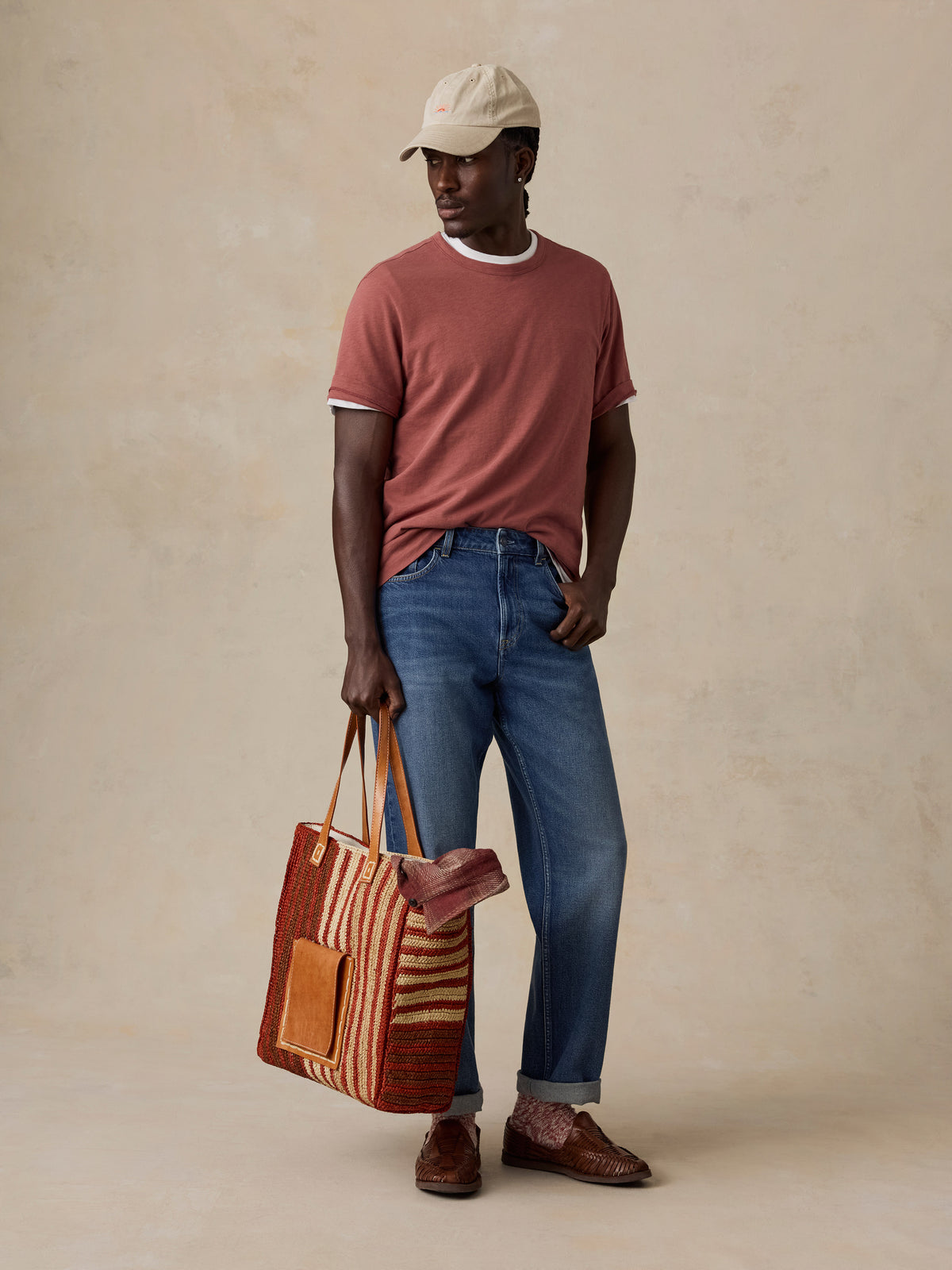 A man wearing a beige baseball cap, a dusty rose ringer t-shirt, blue jeans, and brown leather loafers stands against a textured beige studio backdrop, holding a striped woven tote bag., image 6 of 6