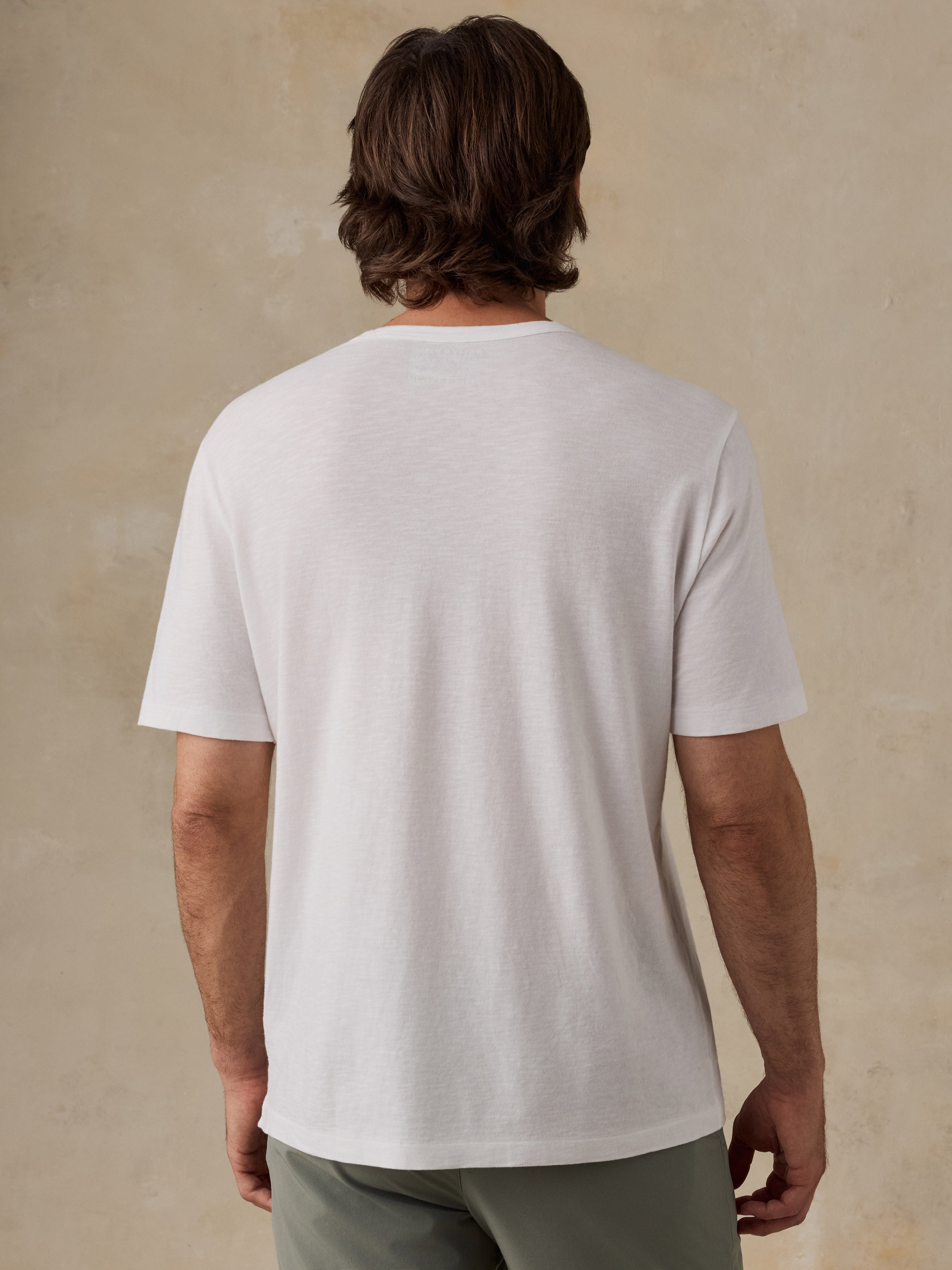 A man viewed from the back wearing a plain white short-sleeve crewneck t-shirt and olive green shorts, standing against a textured beige wall.
