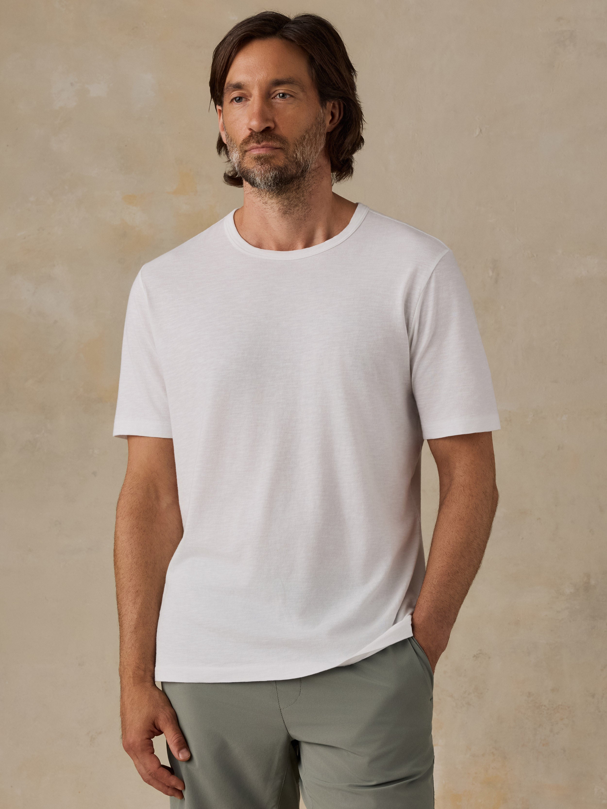 A man wearing a plain white short-sleeve crewneck t-shirt and green shorts, standing against a textured beige wall.