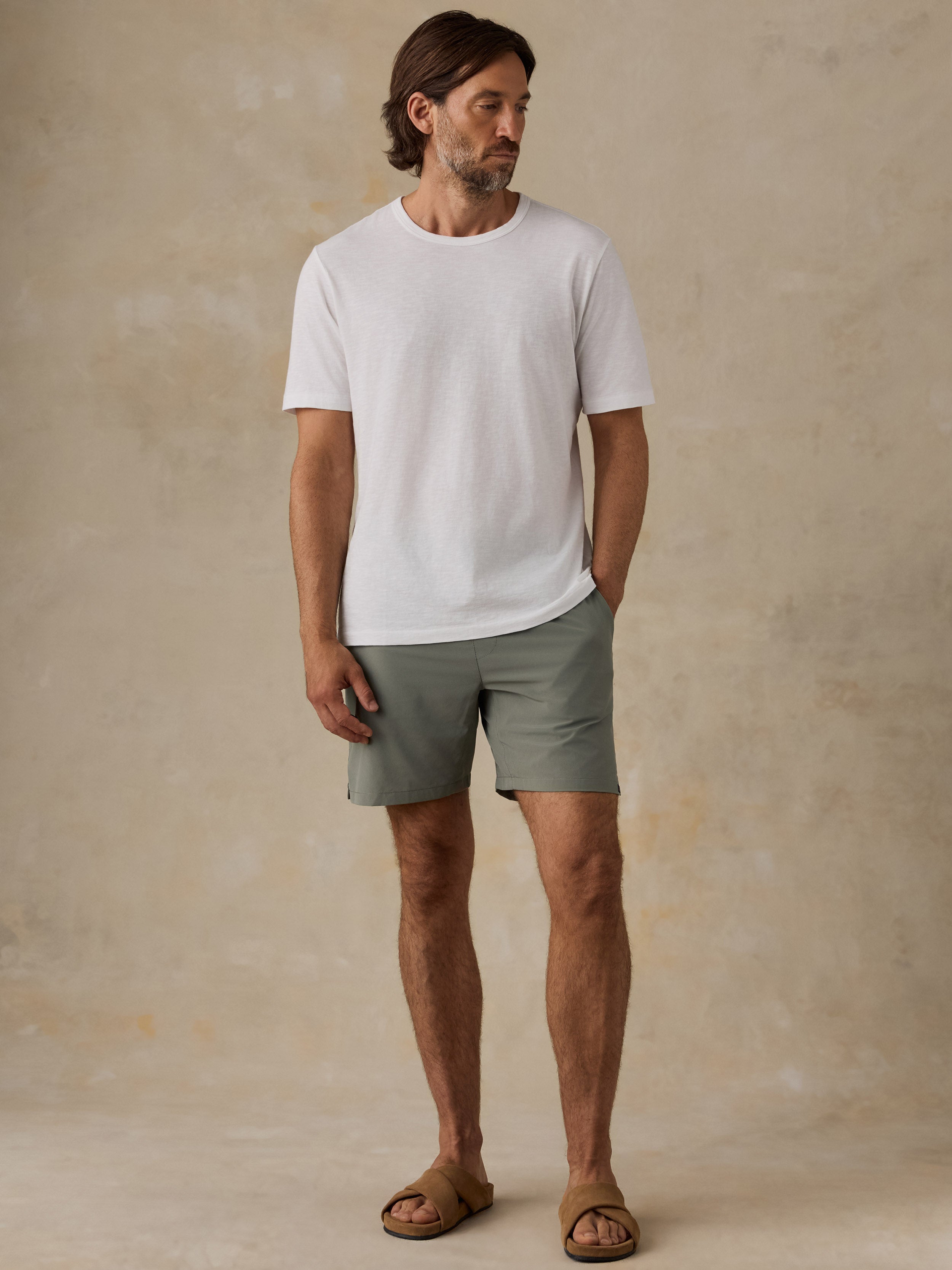 A man wearing a white crew-neck t-shirt, sage green shorts, and brown leather slide sandals, standing against a textured beige studio backdrop.
