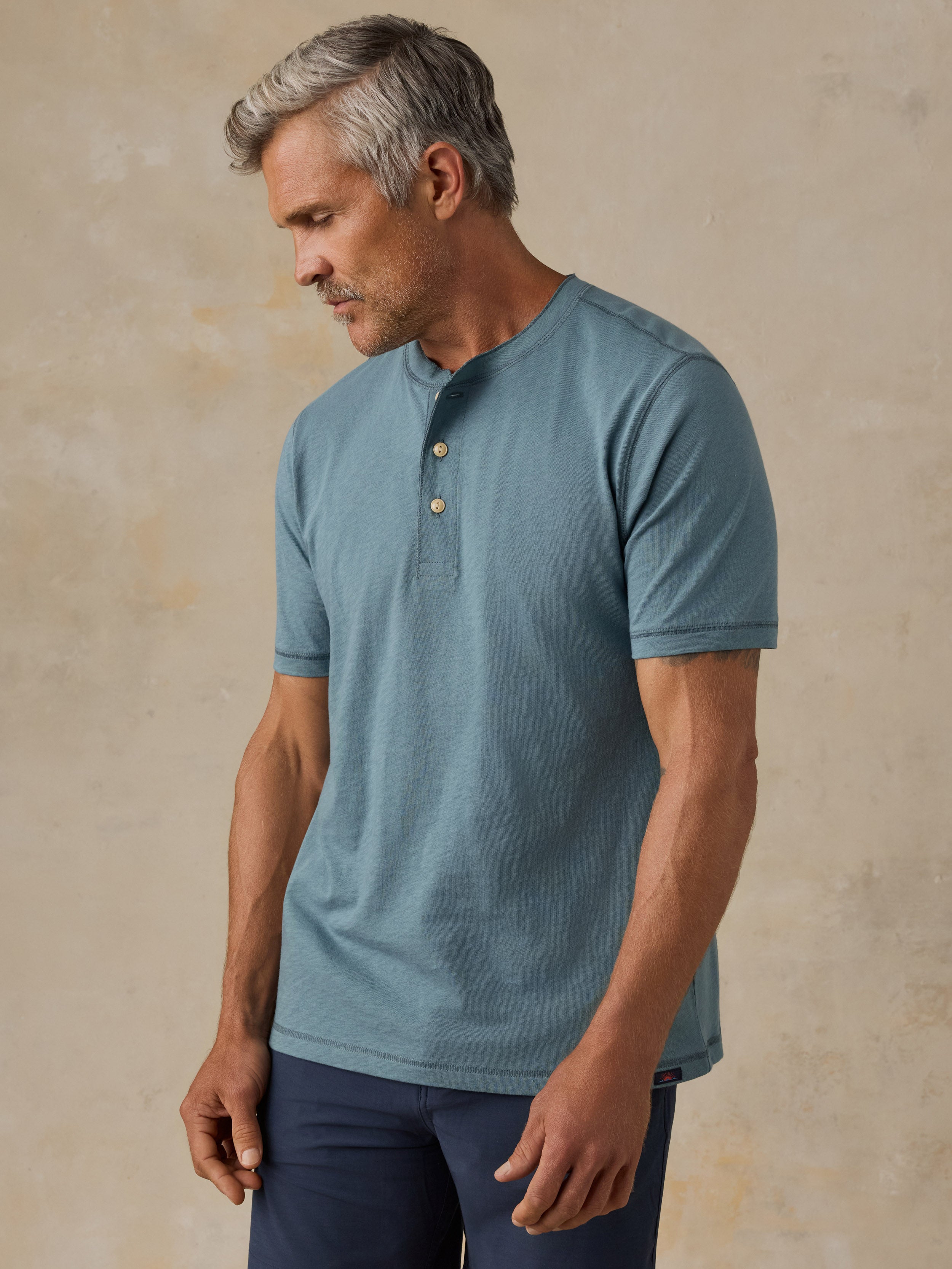 A man wearing a muted blue short-sleeve henley shirt with a three-button placket, standing against a textured beige studio backdrop.