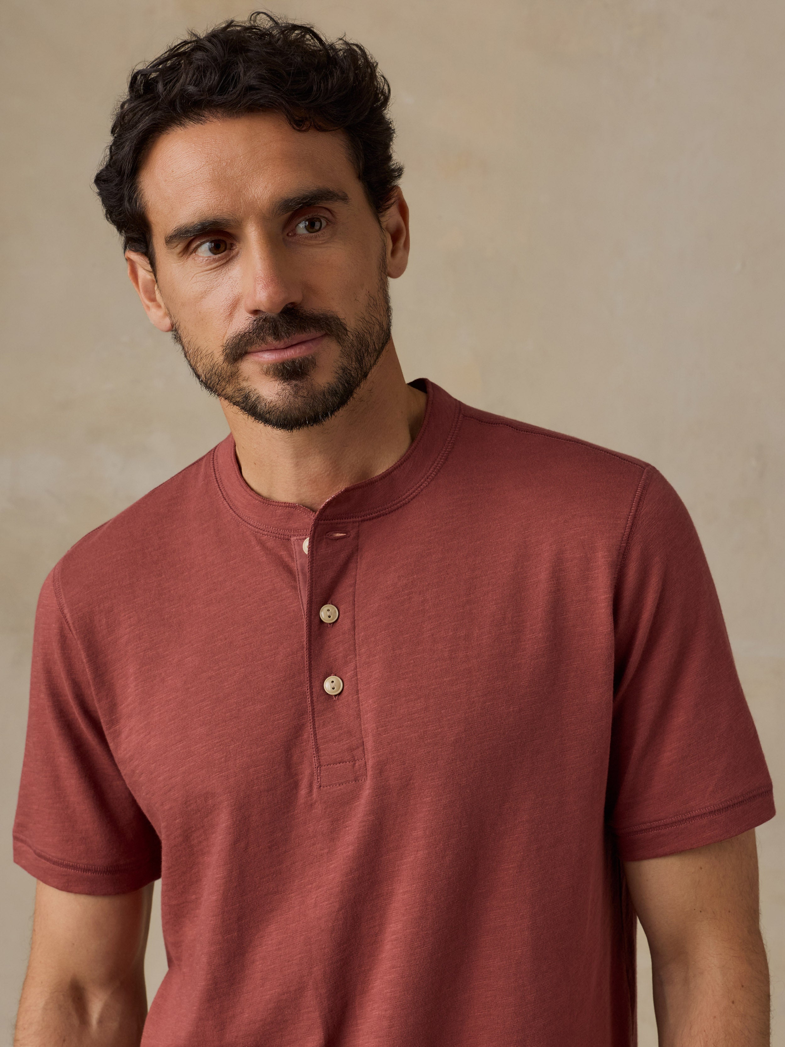 A man wearing a short-sleeve henley shirt in a muted terracotta or dusty rose-red color, standing against a textured beige wall.